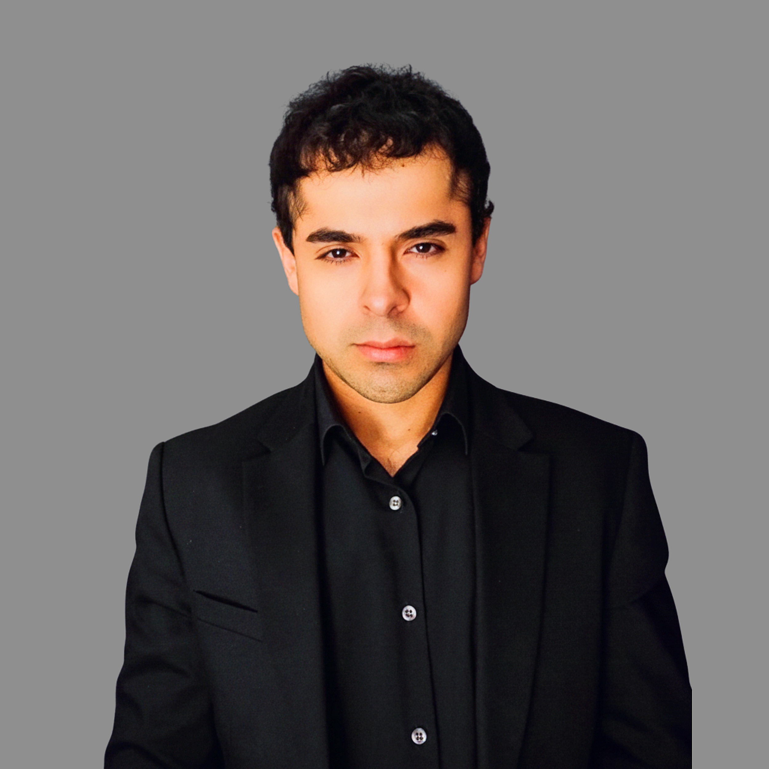 A young man with short dark hair and light skin tone wearing a black suit and black shirt against a plain gray background.