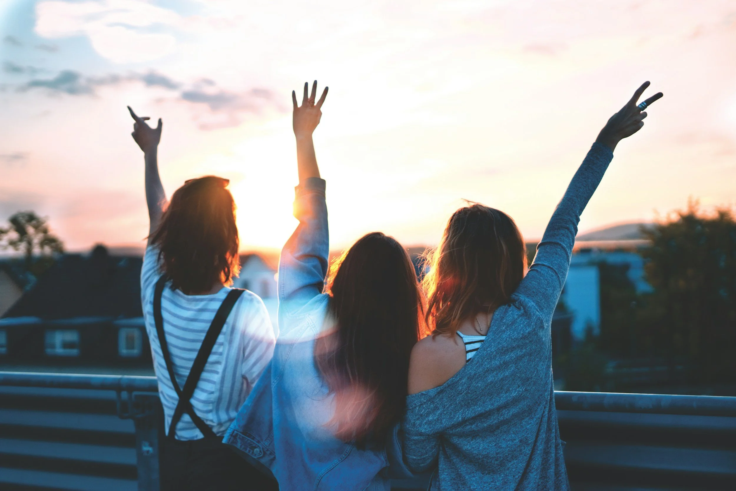 Three young women standing on a balcony during sunset with their backs to the camera, raising their arms in celebration.