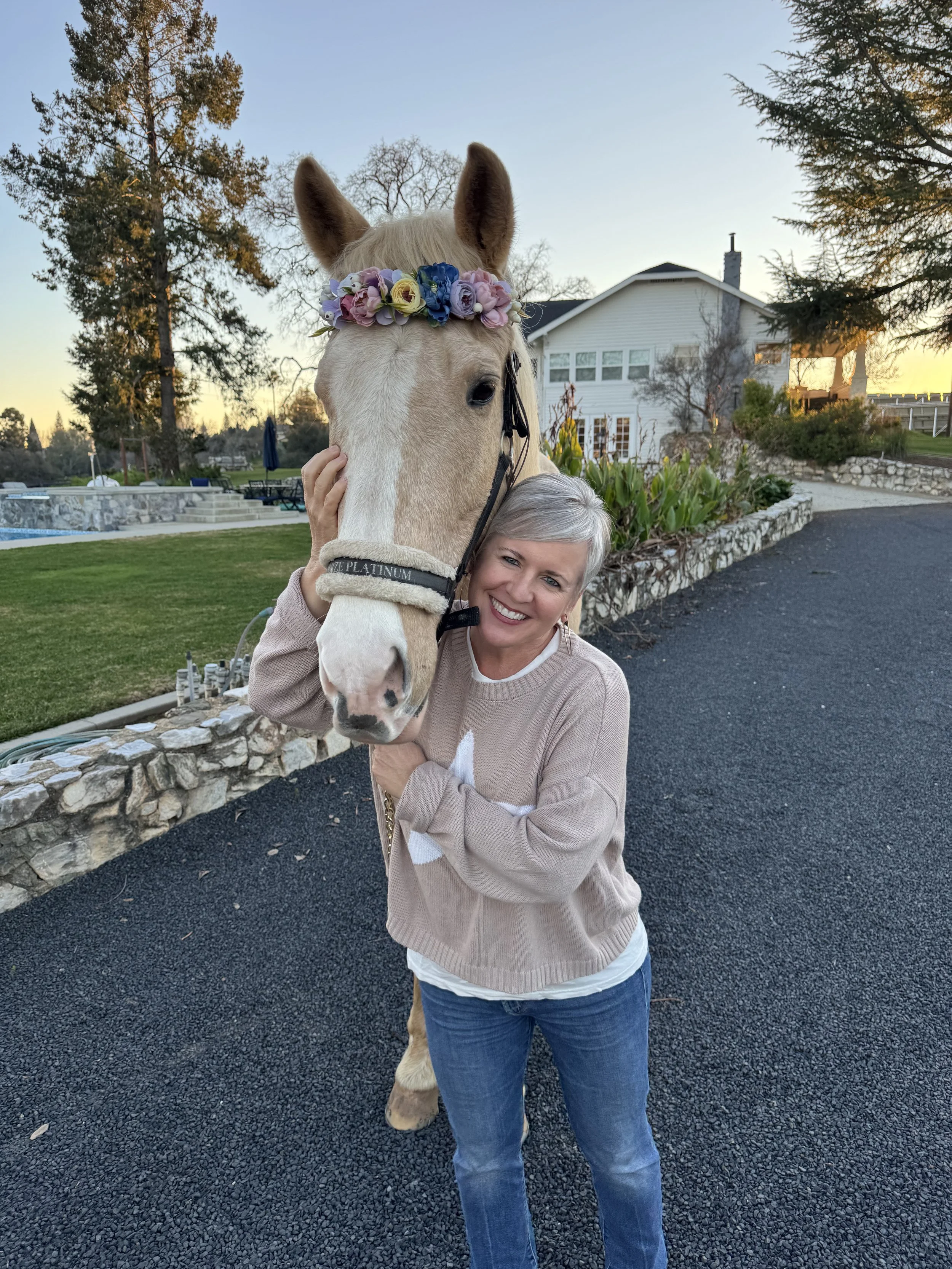 Smiling woman hugging a light-colored horse wearing a floral crown in a garden setting at sunset.