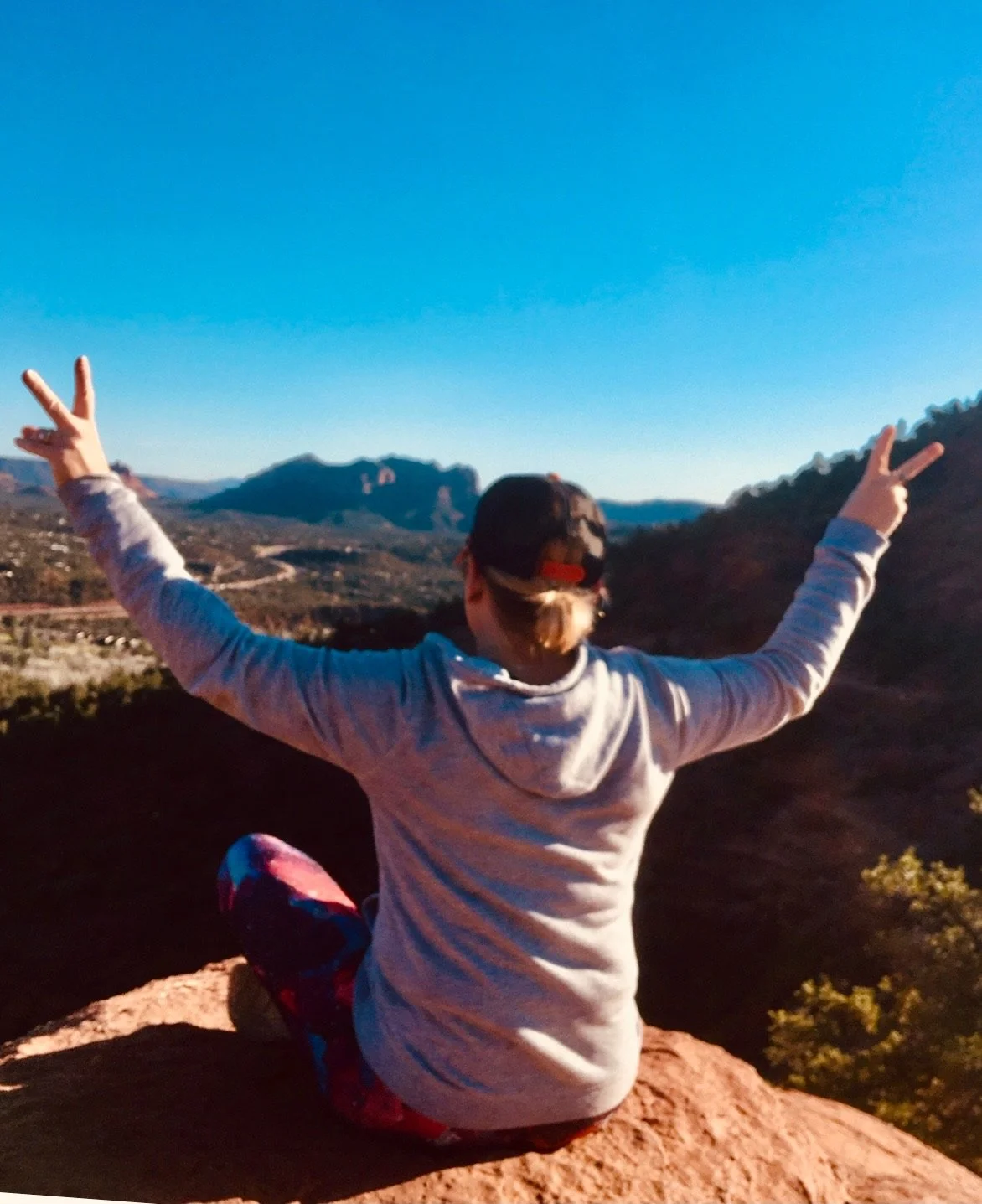 Person sitting on rock at scenic overlook with arms raised in victory pose, mountain landscape in background under clear blue sky.