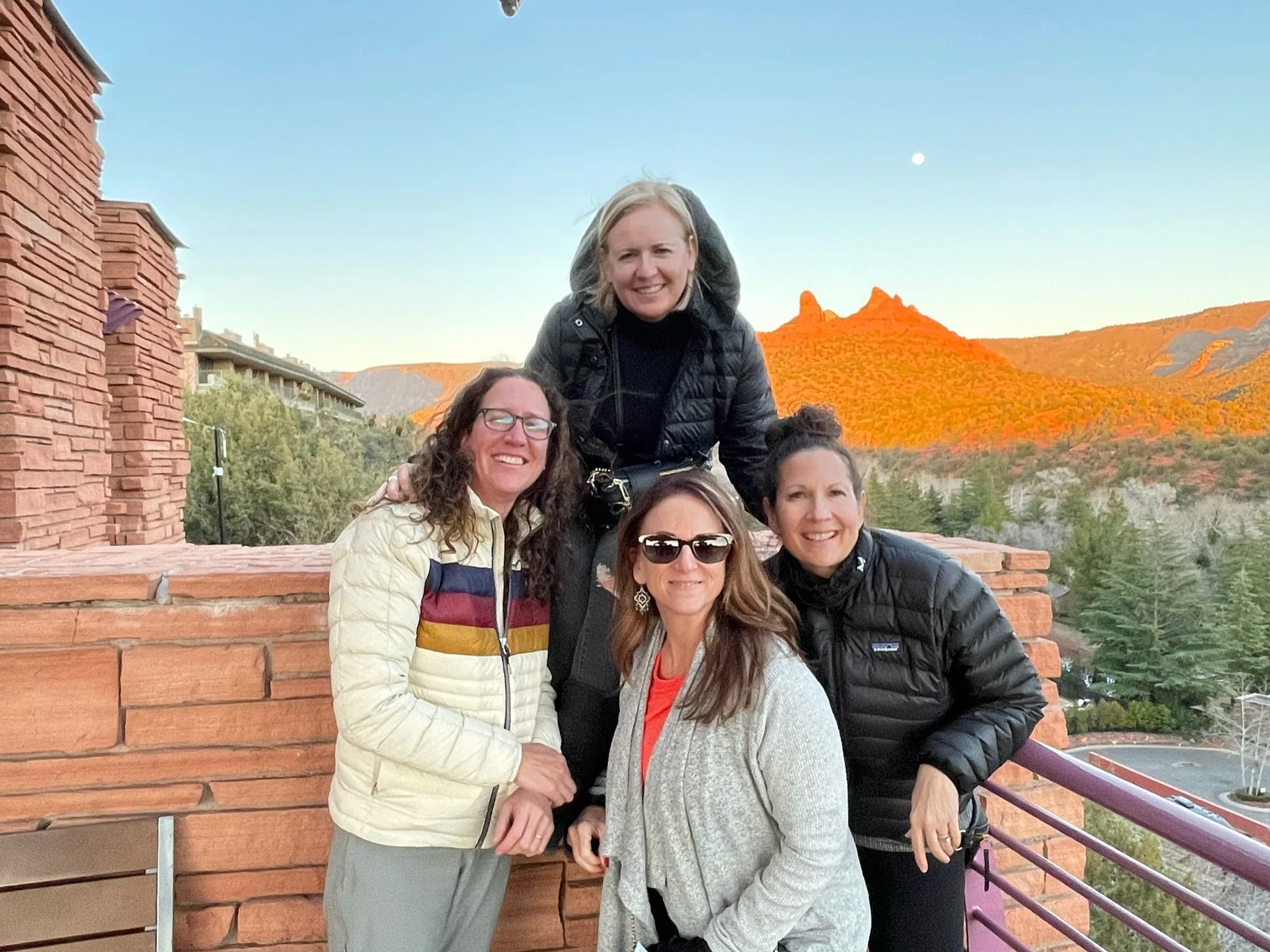 Four women smiling on a scenic outdoor balcony with red rock formations and trees in the background during sunset or sunrise.