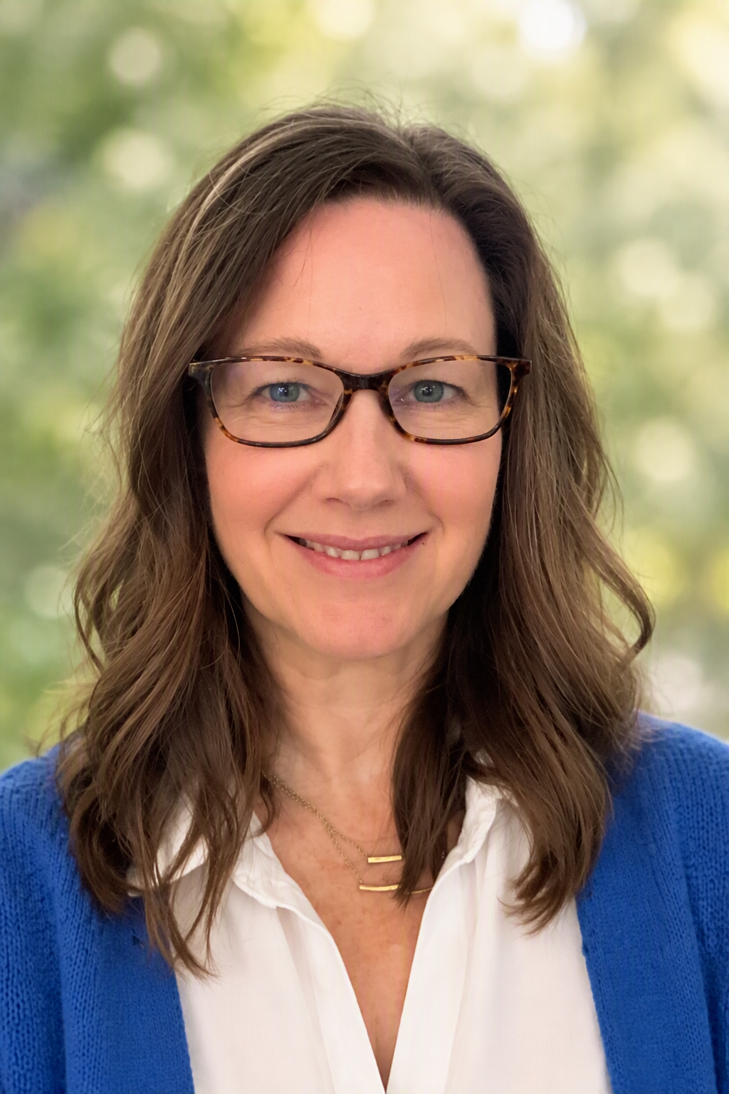 Portrait of a woman with shoulder-length brown wavy hair wearing glasses, a white blouse, and a blue cardigan, smiling against a blurred outdoor background.