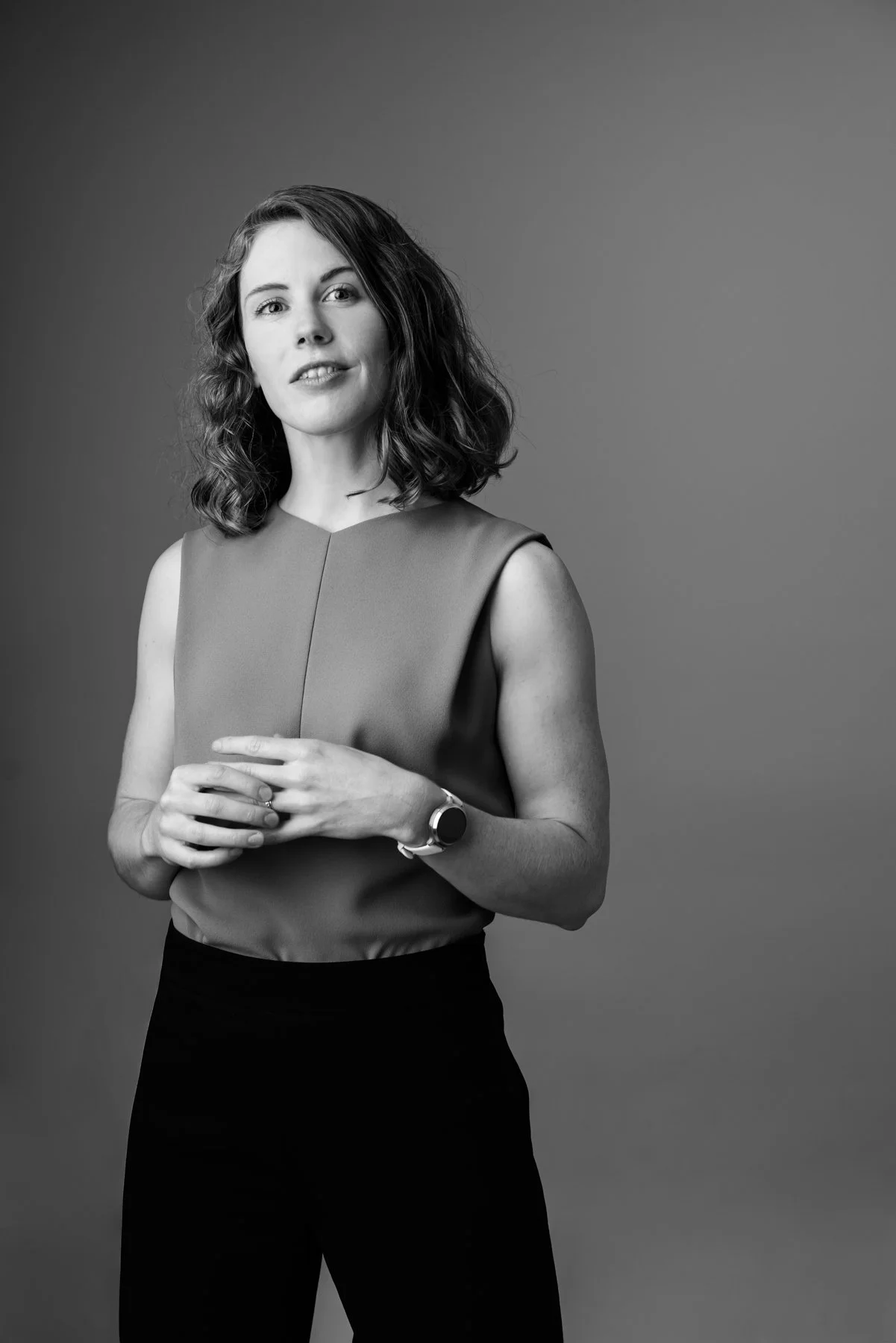 A black and white portrait of a woman with shoulder-length wavy hair, wearing a sleeveless top and dark pants, standing against a plain background.
