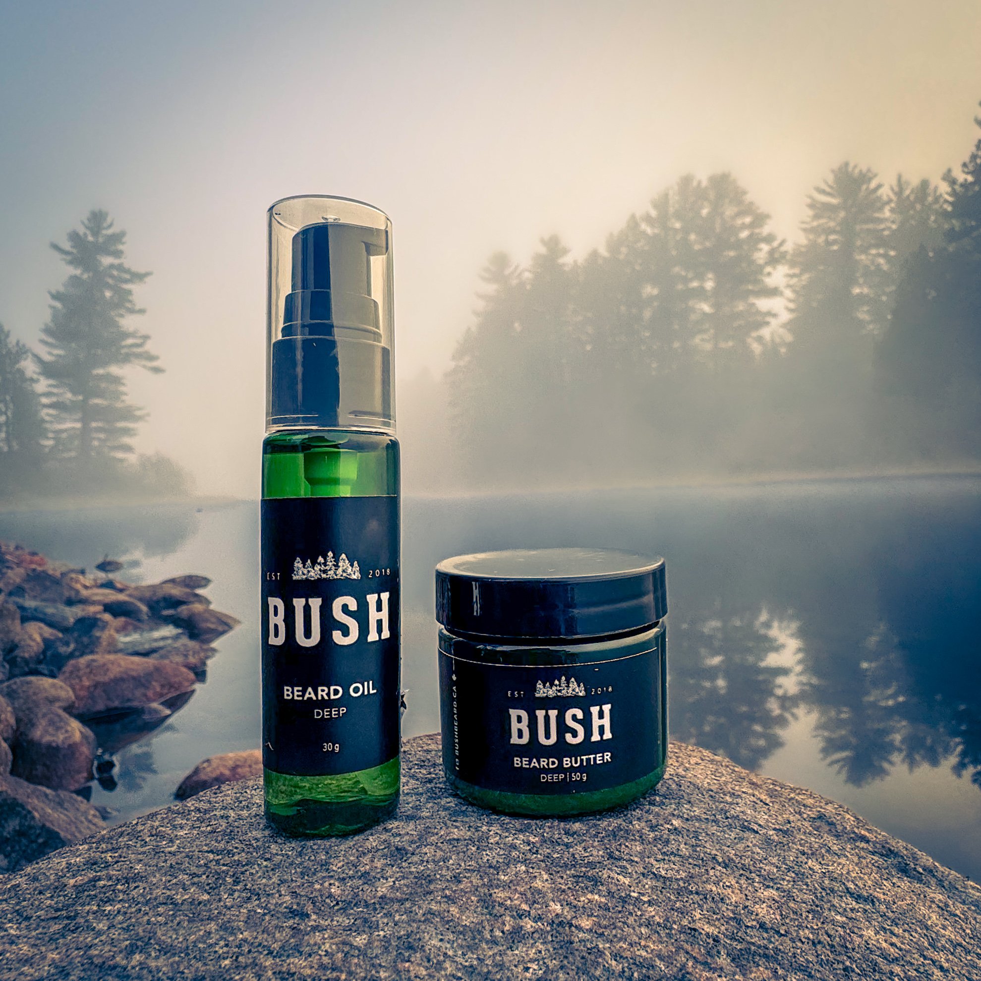 Bash beard oil and beard butter products on a rock by a foggy lake with trees in the background.