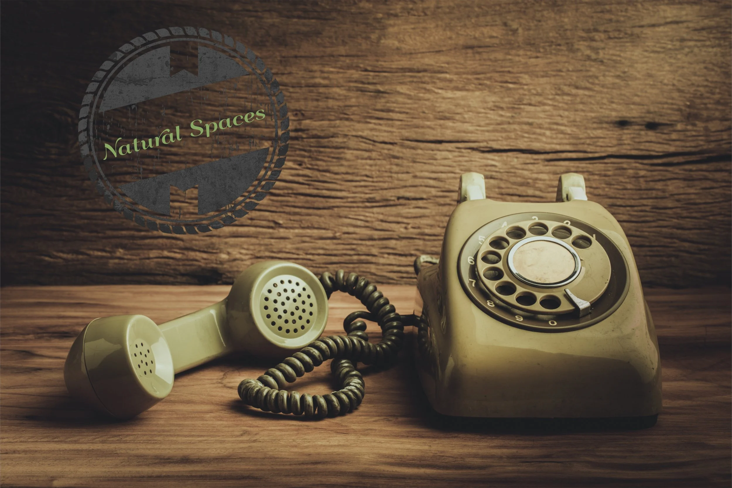 A vintage beige rotary telephone on a wooden surface with a wooden backdrop, with a green logo that reads 'Natural Spaces' in the upper left corner.