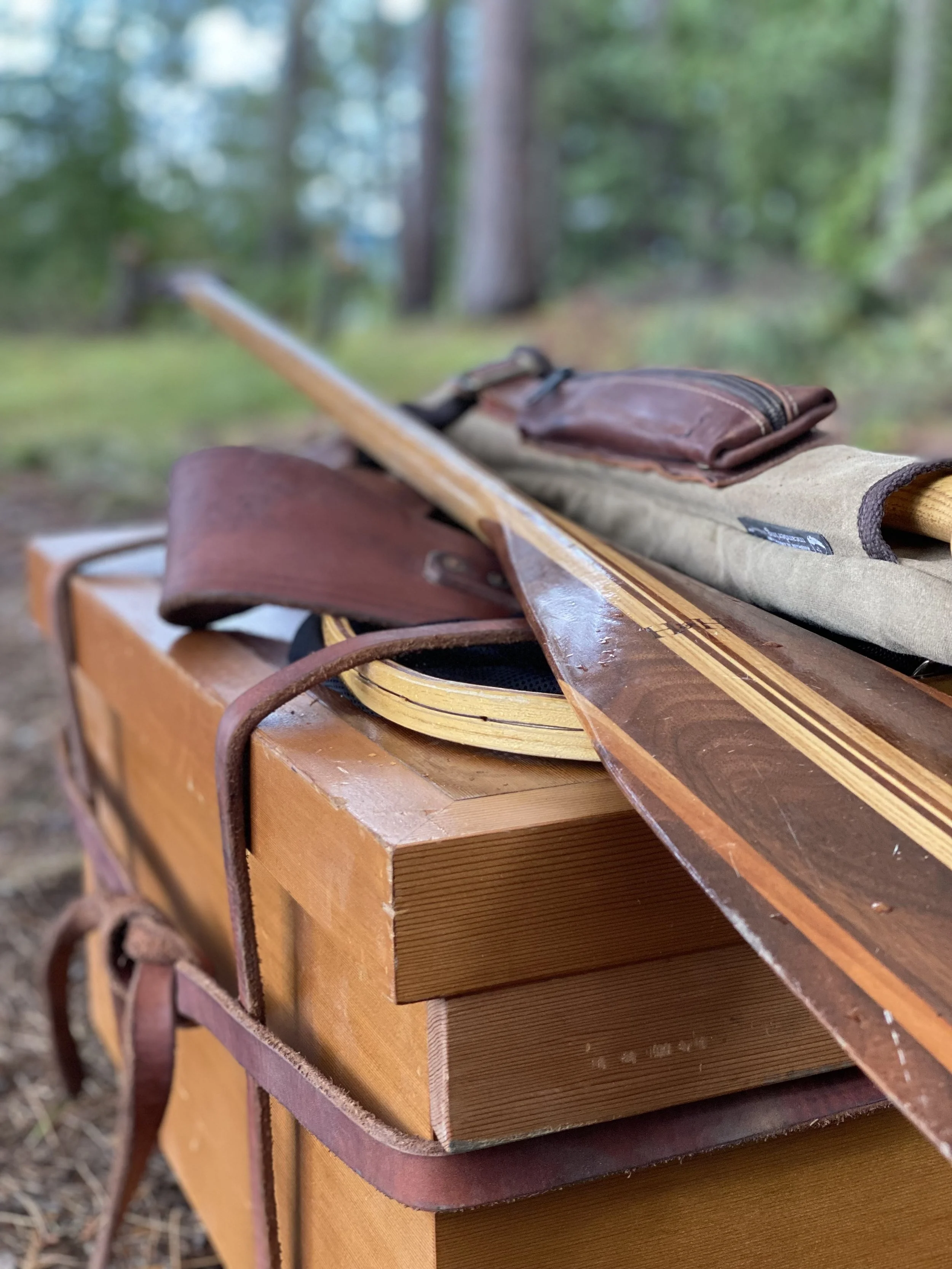 A wooden box with a wooden paddle, a leather strap, and a rolled-up strap on top, with trees in the background.