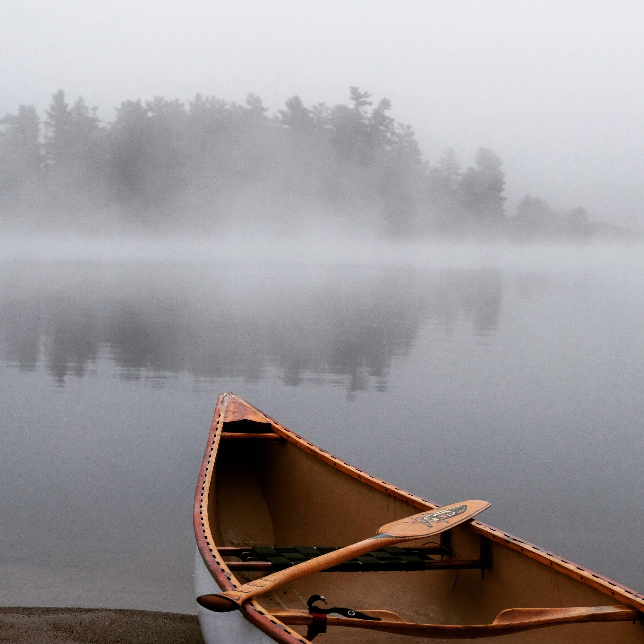 A canoe resting on the shore of a calm lake with foggy trees in the background.