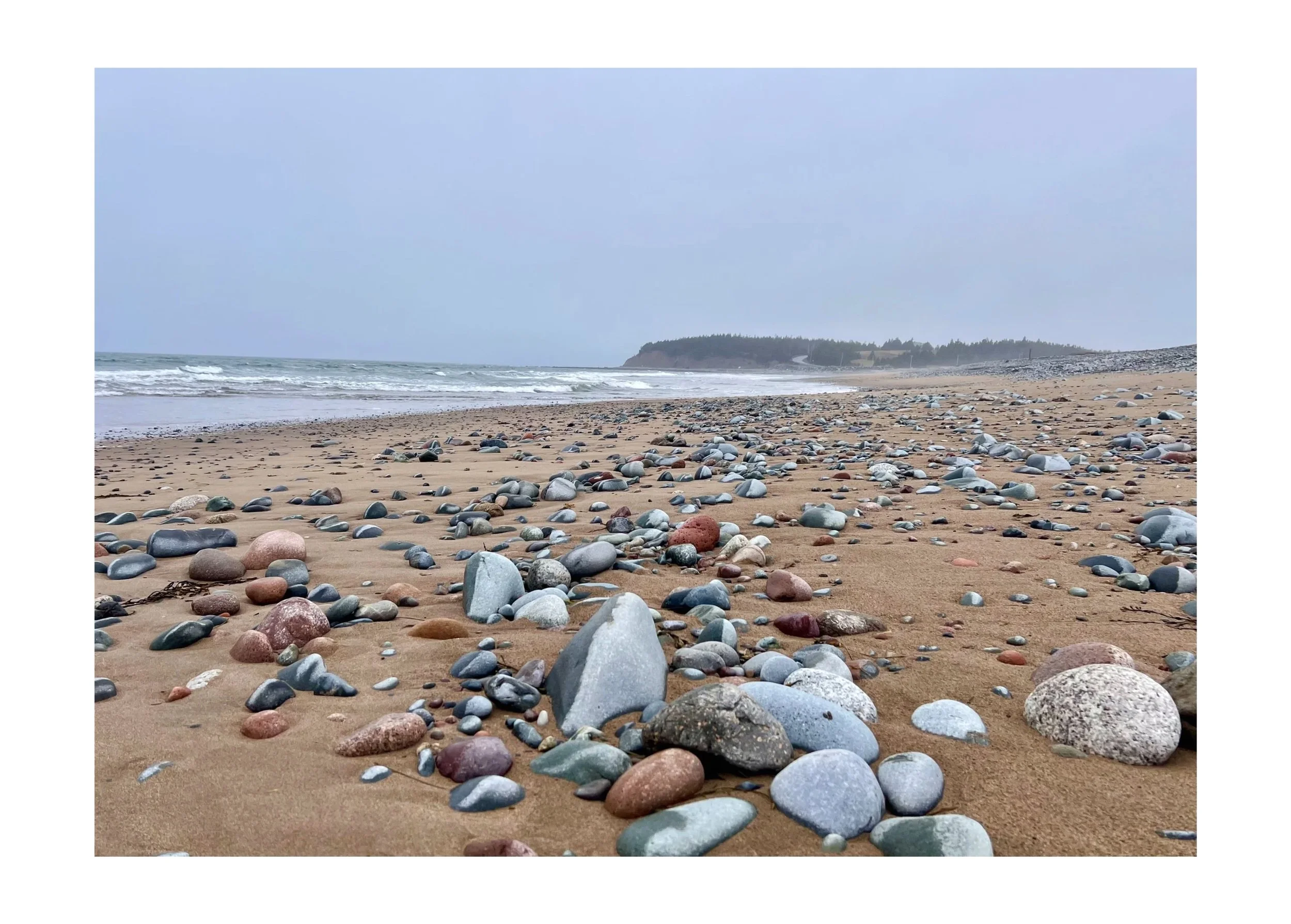 A sandy beach with smooth, colorful pebbles scattered across the shore, gentle waves approaching, and a distant tree-lined headland under a cloudy sky.