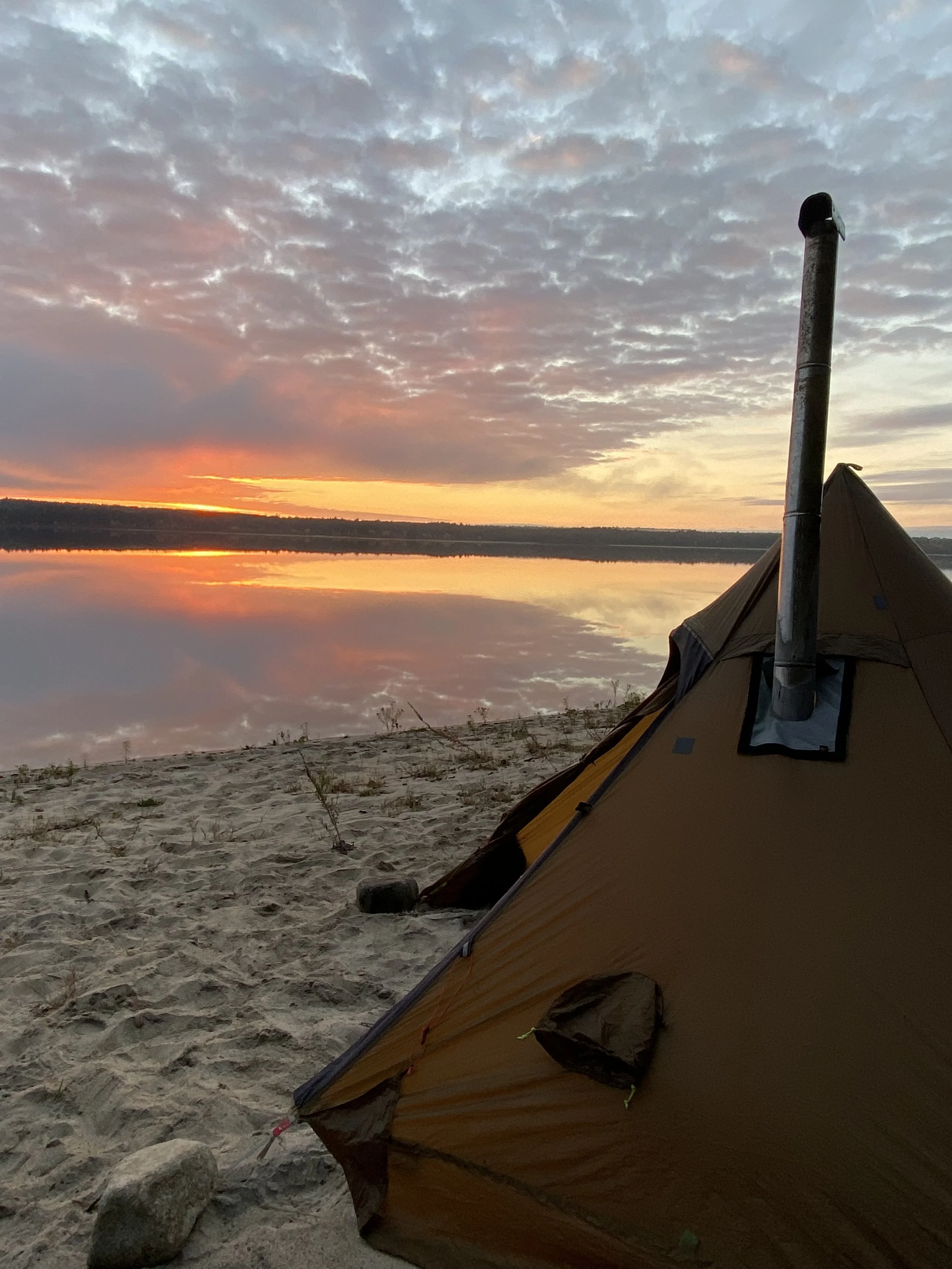 Tent pitched on sandy shore beside a calm lake during sunset, with colorful clouds reflected in the water and a small rock in the foreground.