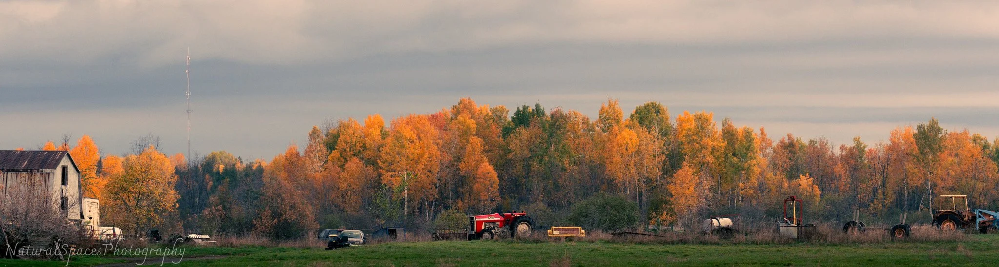 A rural scene with an old barn, tractors, and farm equipment in front of a treeline with autumn-colored leaves, under a cloudy sky.