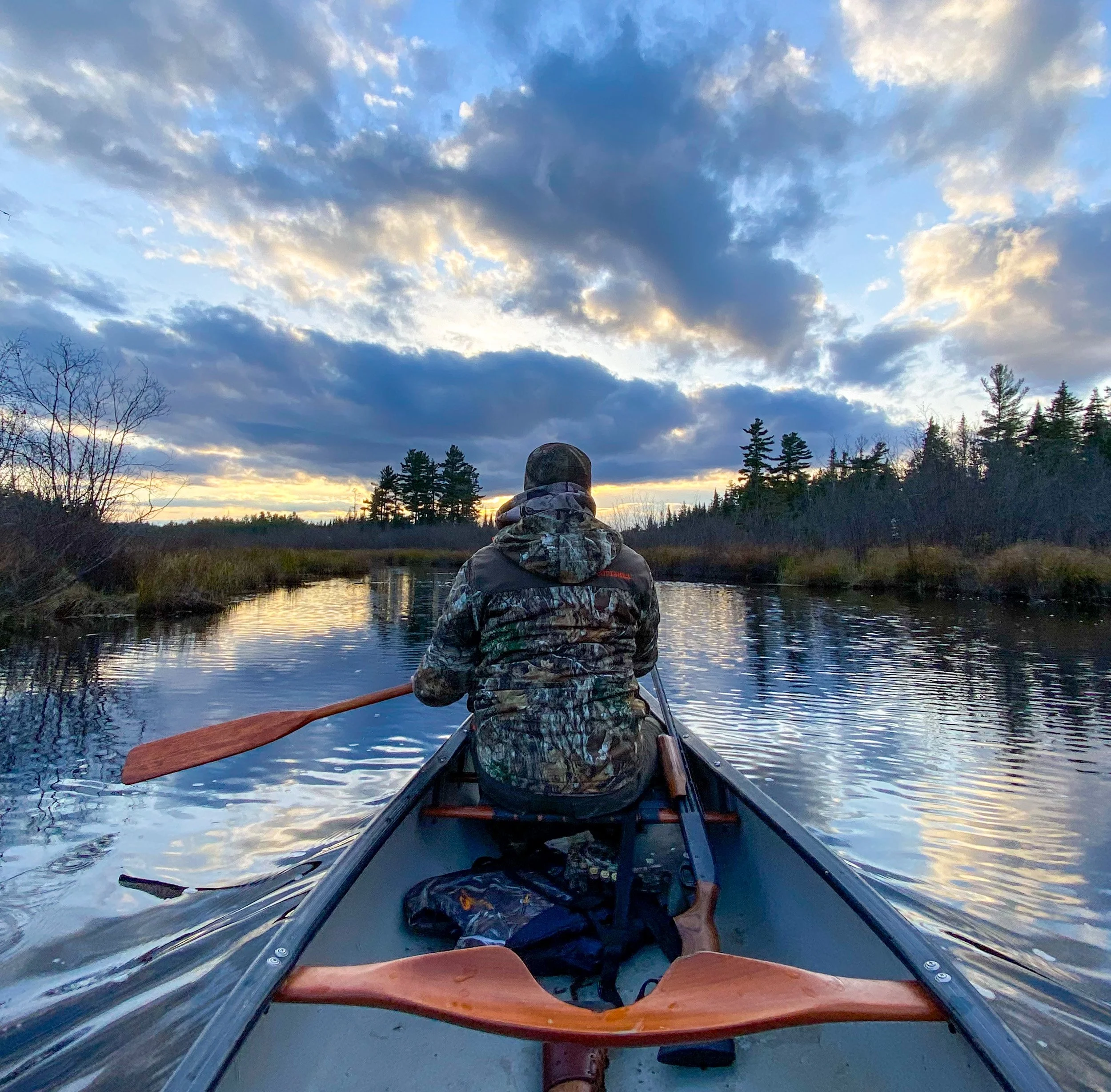 A person wearing camouflage clothing and a hat is sitting in a canoe, paddling on a river during sunset or sunrise surrounded by trees and clouds.