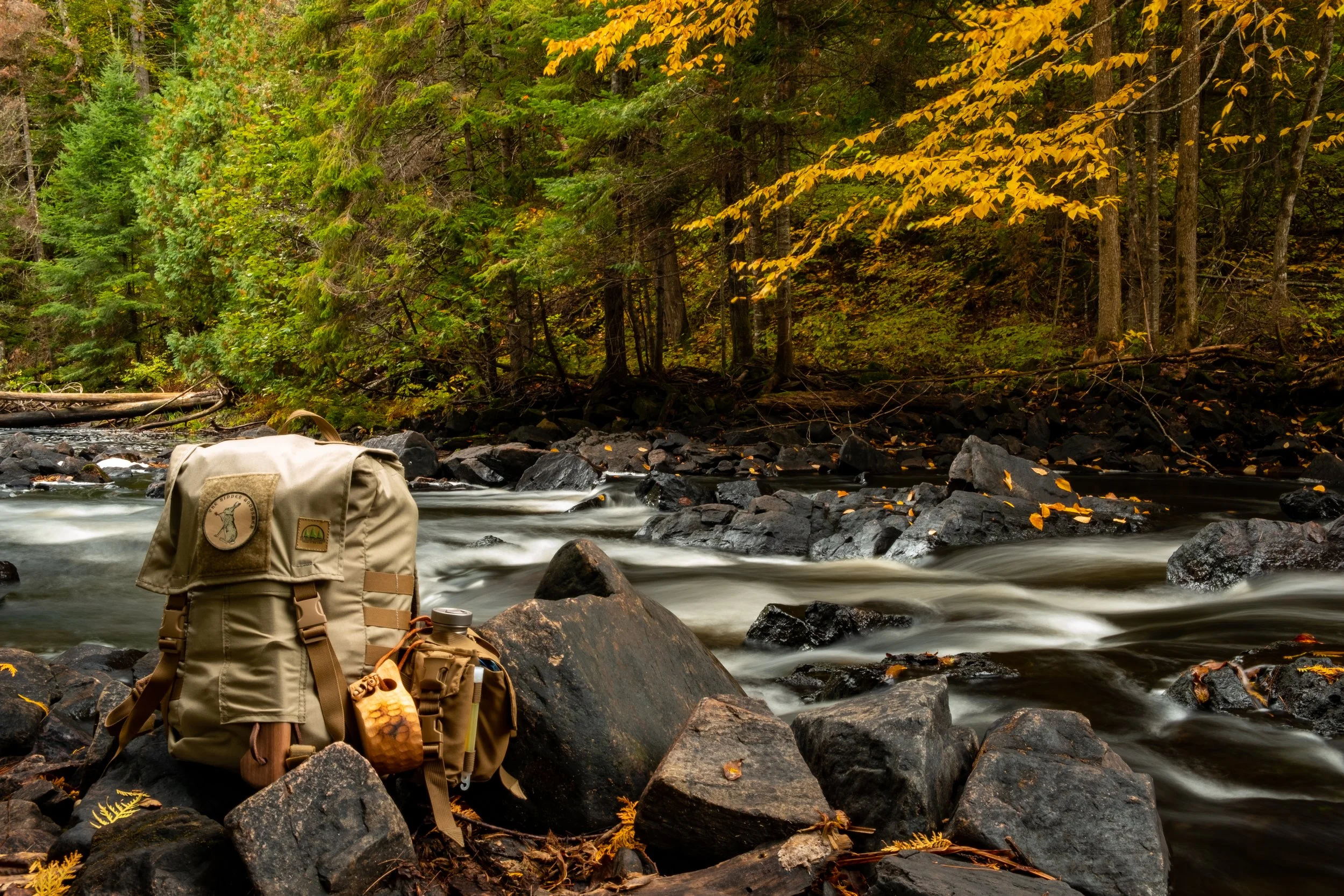 A beige backpack with patches, beside a water bottle, on rocks by a flowing river with a forested background showing trees with green and yellow leaves.