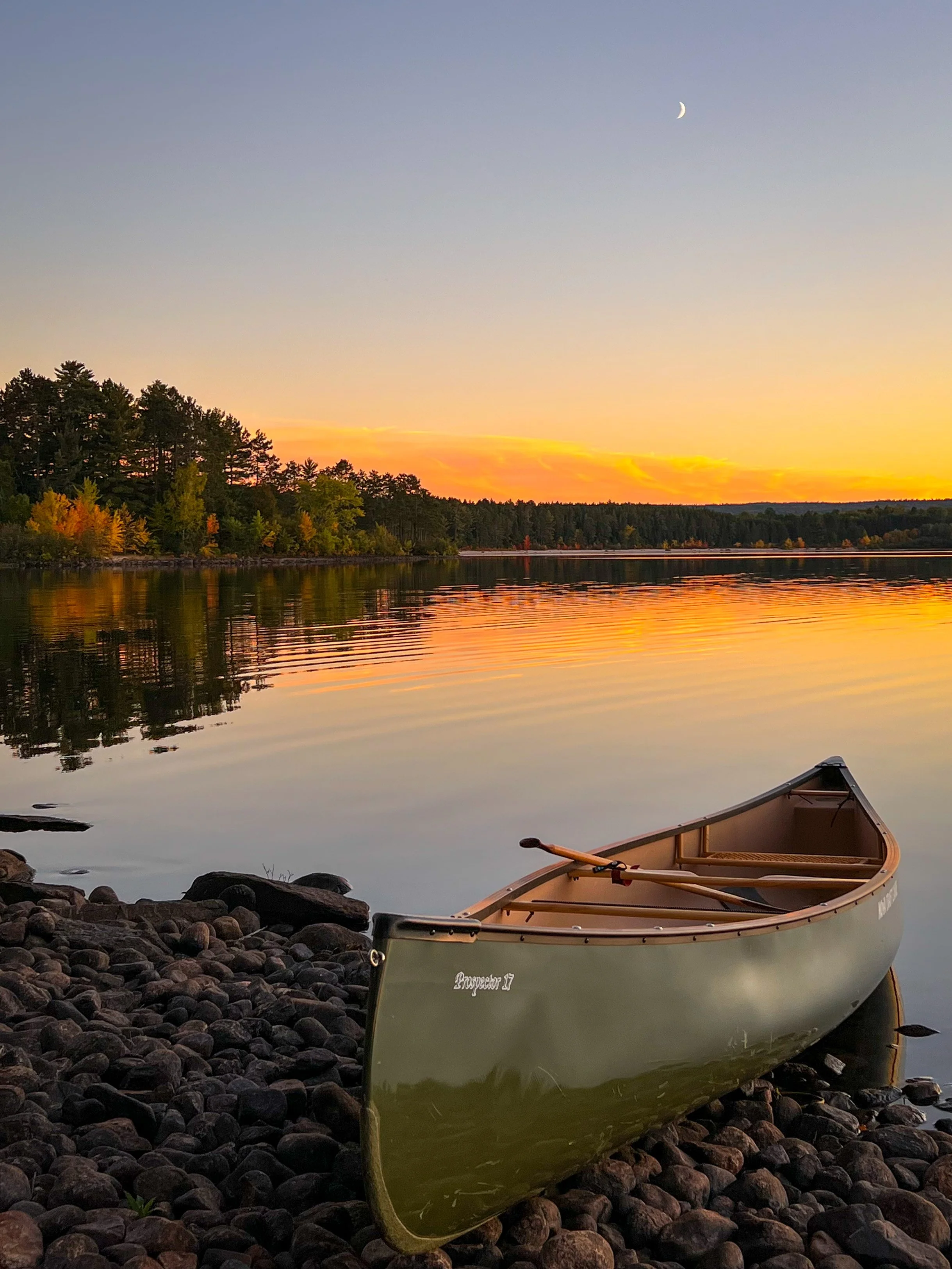 A canoe resting on a rocky shoreline at sunset, with calm water reflecting the colorful sky