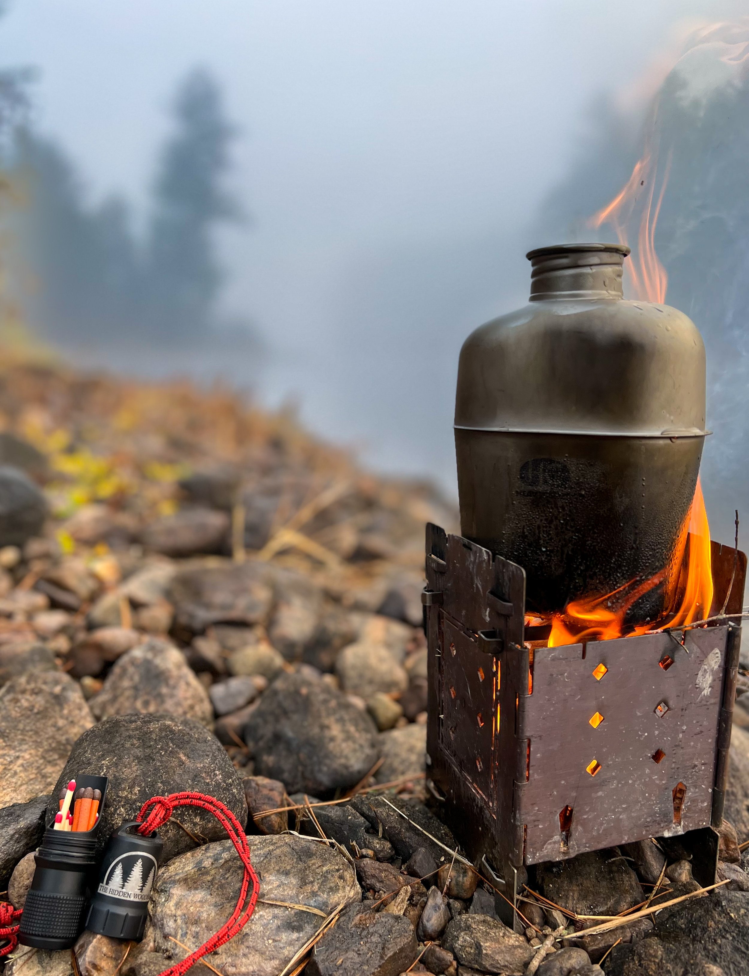 A gray camping stove with a large metal container on top, producing flames, set on rocky terrain with blurred trees and mist in the background. A small black flashlight with a red lanyard and a pack of matches are on the rocks nearby.