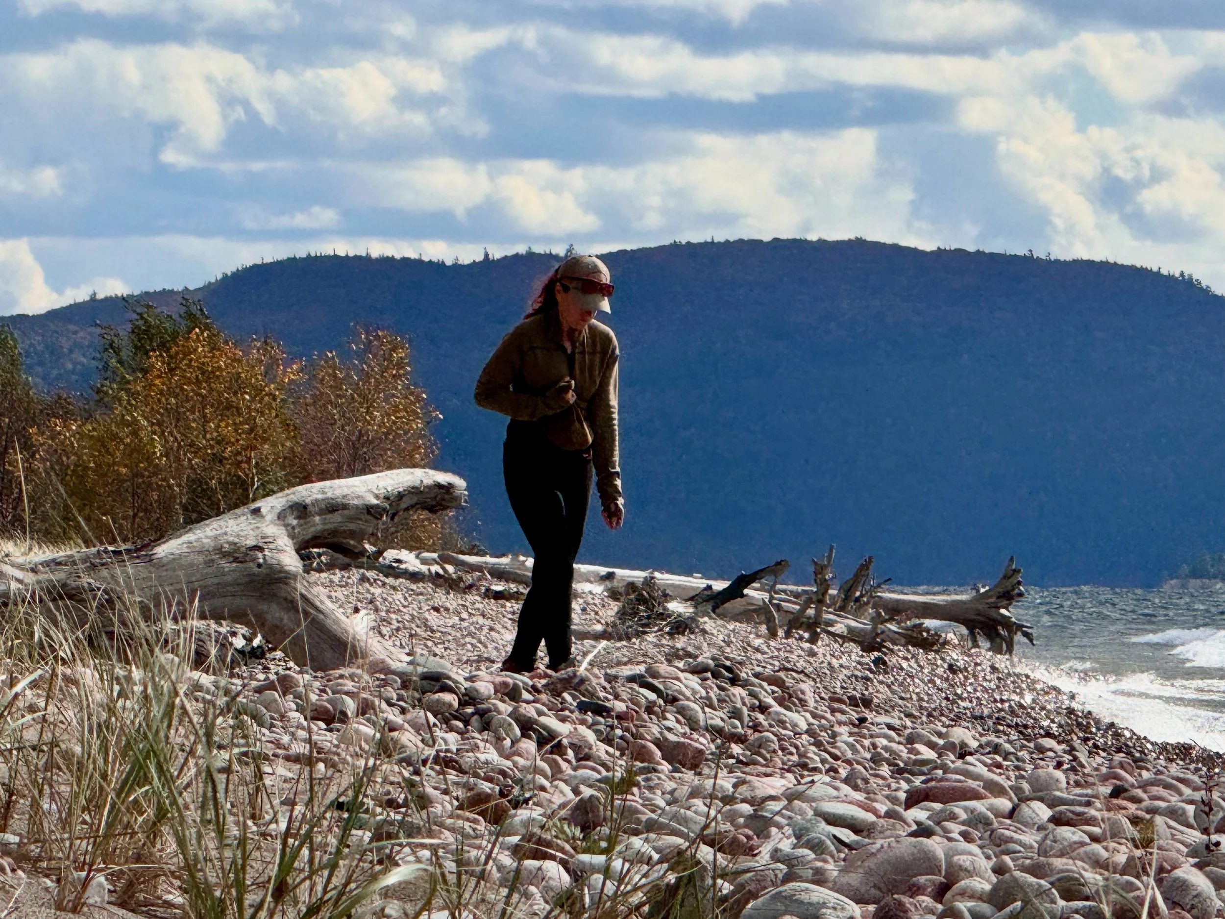 A person in outdoor gear walking along a rocky beach with driftwood, trees, mountains, and cloudy sky in the background.
