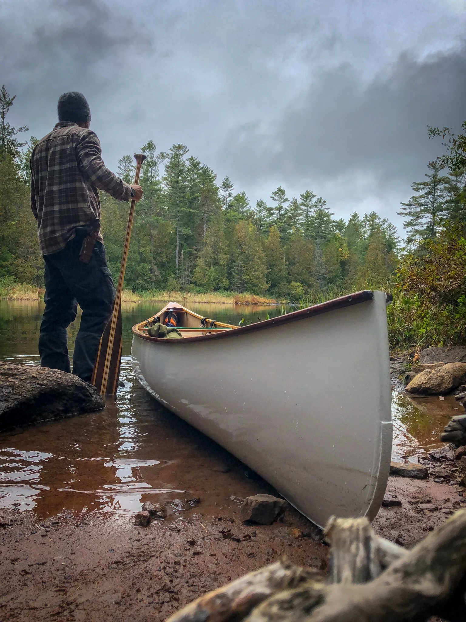 A person standing at the edge of a river with a paddle, preparing to launch a canoe into the water, surrounded by trees and a cloudy sky.