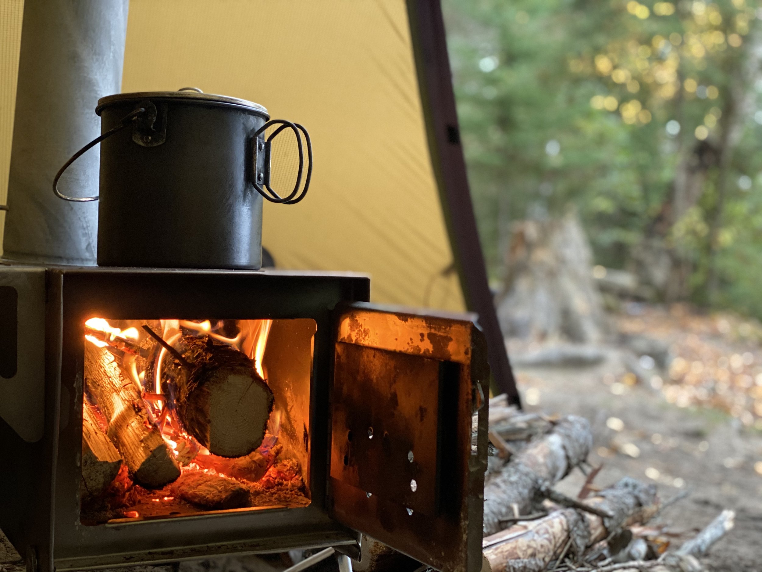 A camping scene with a small wood-burning stove with a pot on top, inside a tent in a forest setting.