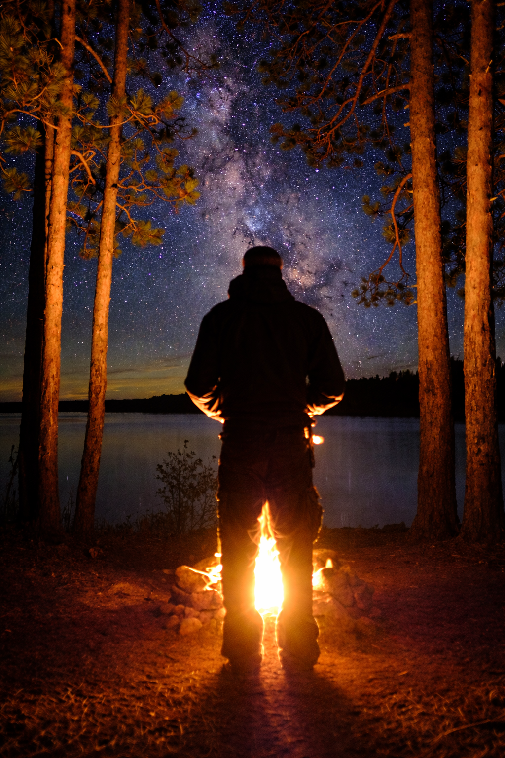 A person standing near a campfire by a lake, gazing at a star-filled night sky 
