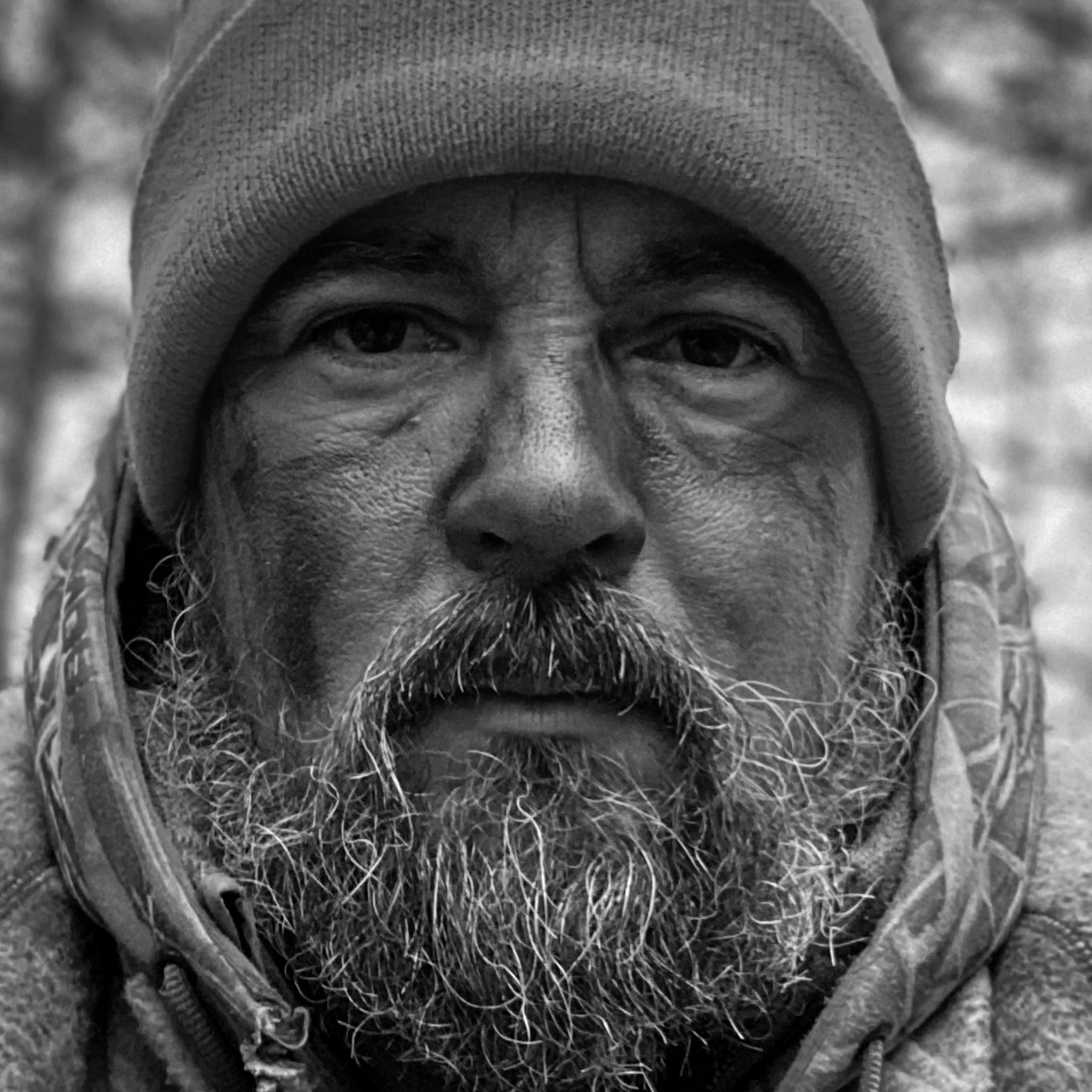 Close-up black-and-white photo of a bearded man wearing a knit cap and a hoodie.
