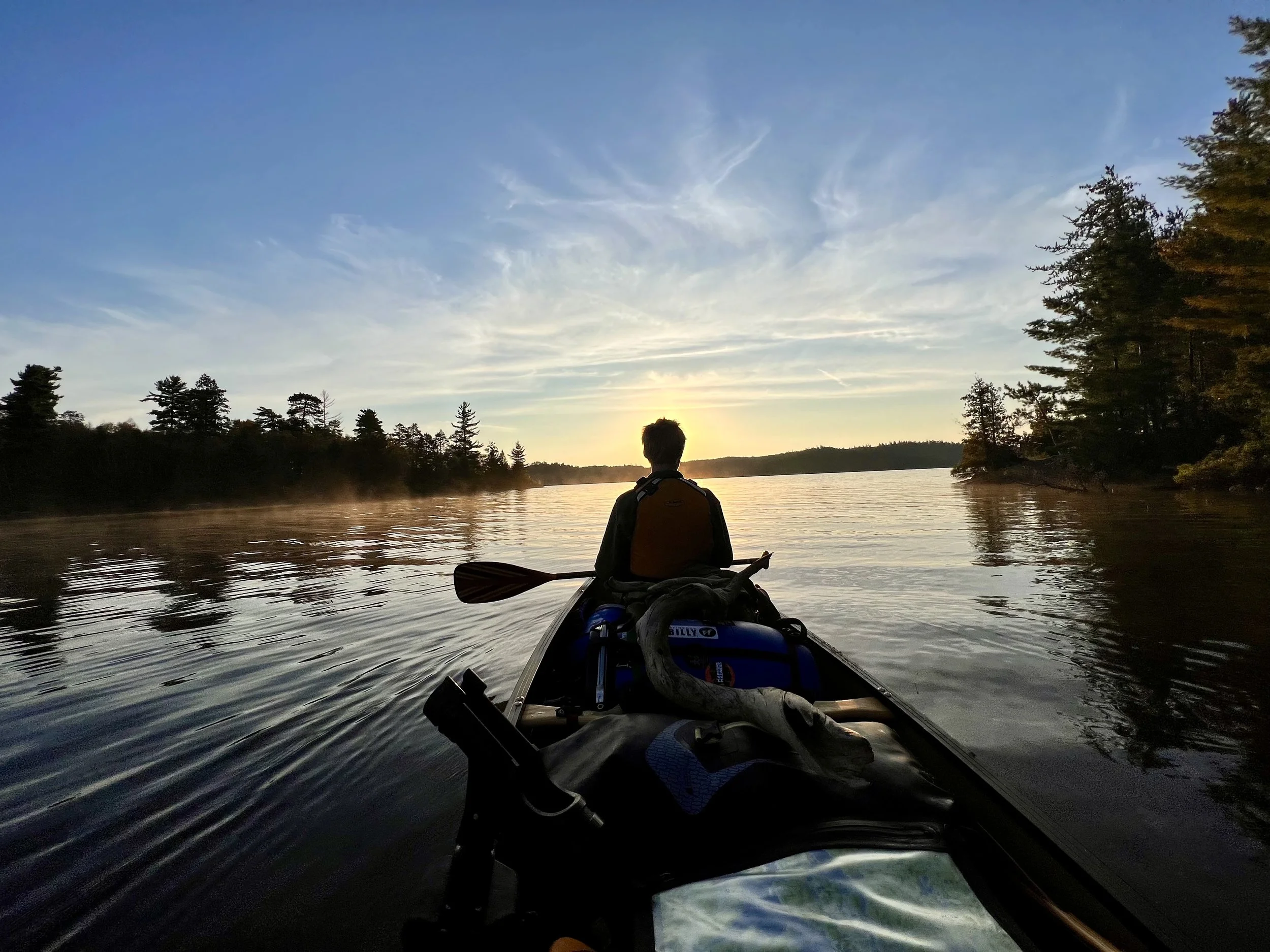 A person kayaking on a calm river at sunset with trees on both sides.