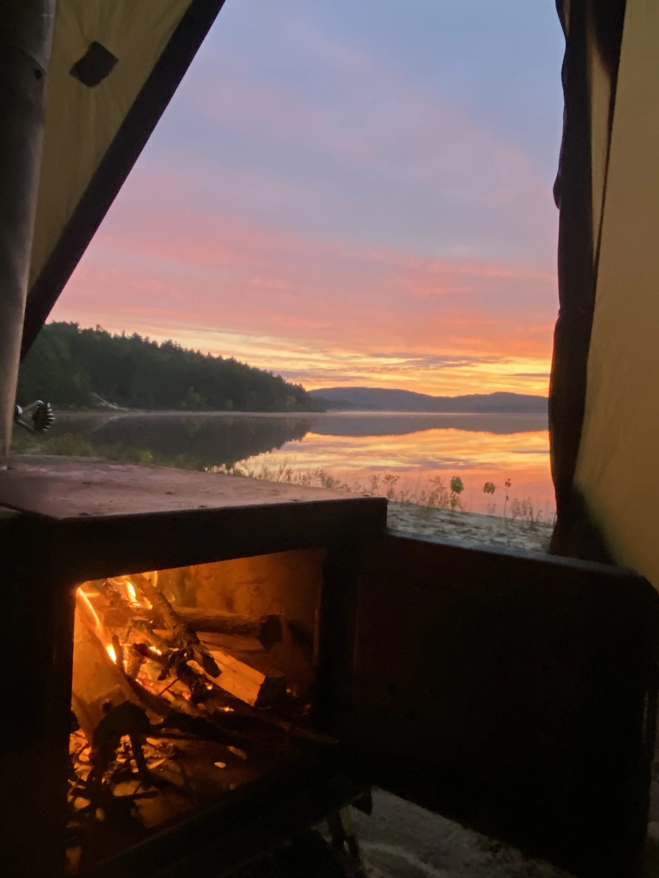 Sunset over a lake viewed from inside a tent with a small campfire.