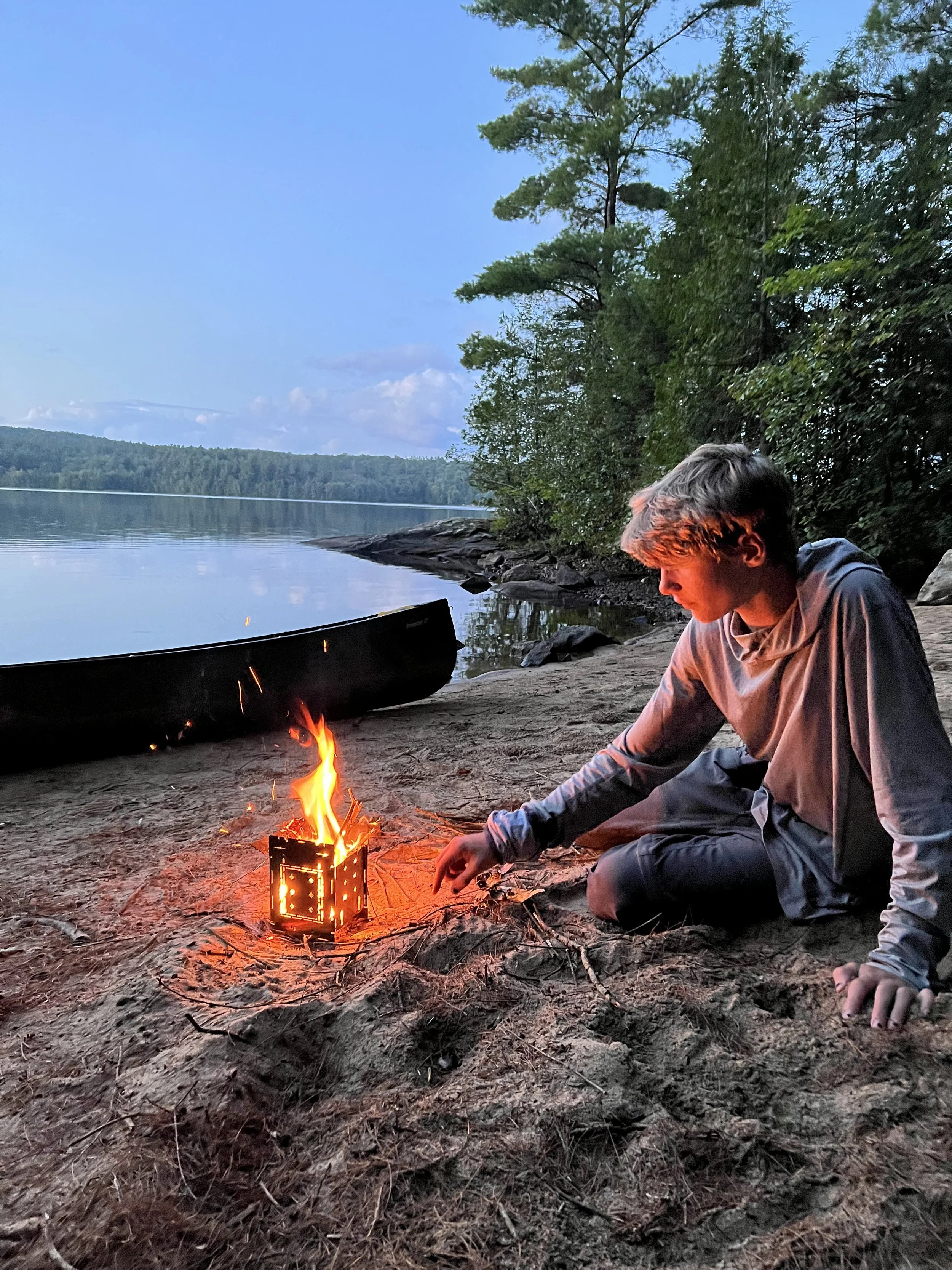A boy outdoors sitting on sandy ground near a body of water, tending to a small campfire with a metal fire pit. Behind him are trees and a canoe on the shore.