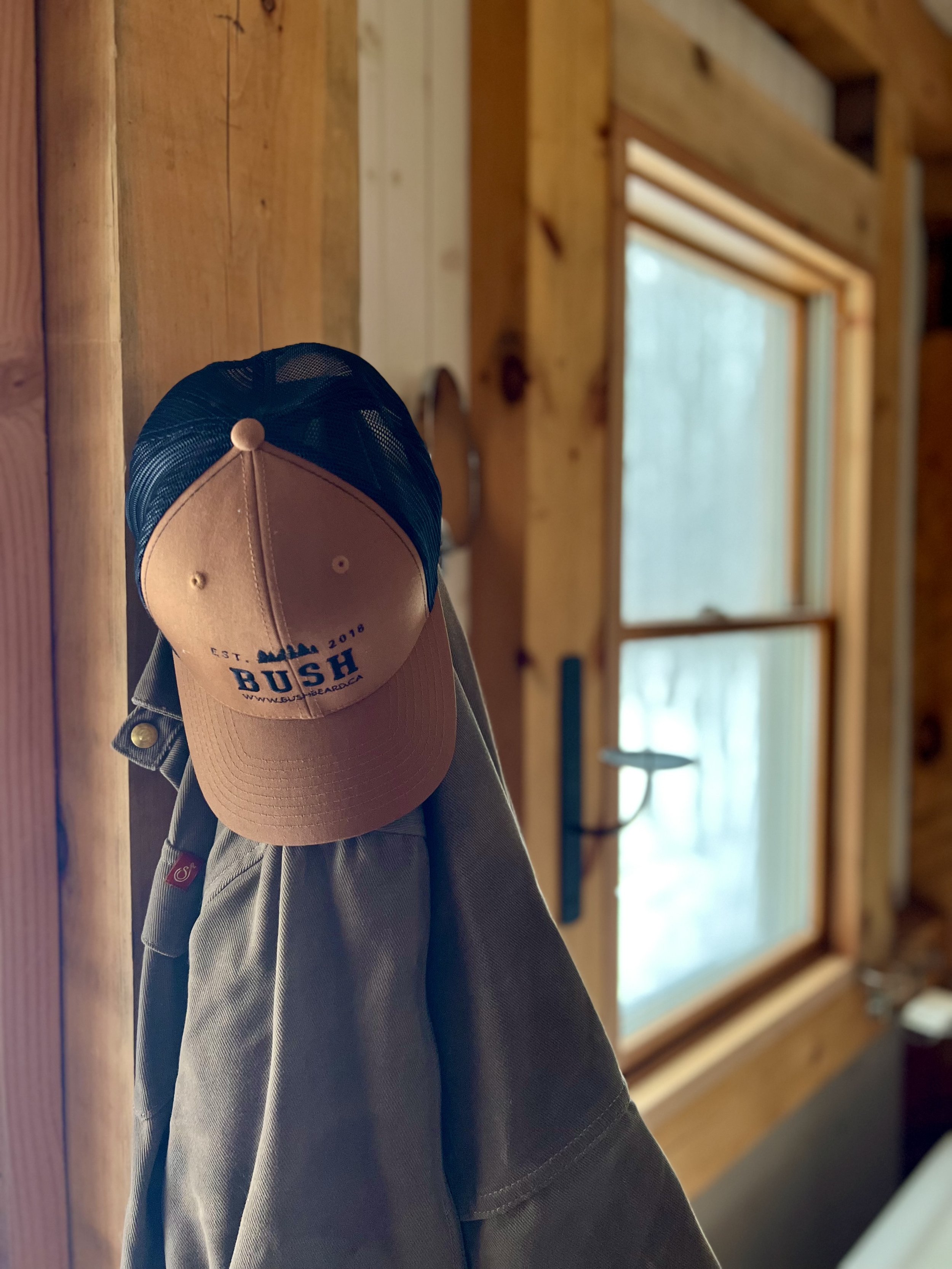 A tan mesh BUSH ball cap hanging on a hook with a tan jacket below it, on a wooden wall in front of a window with light coming through.