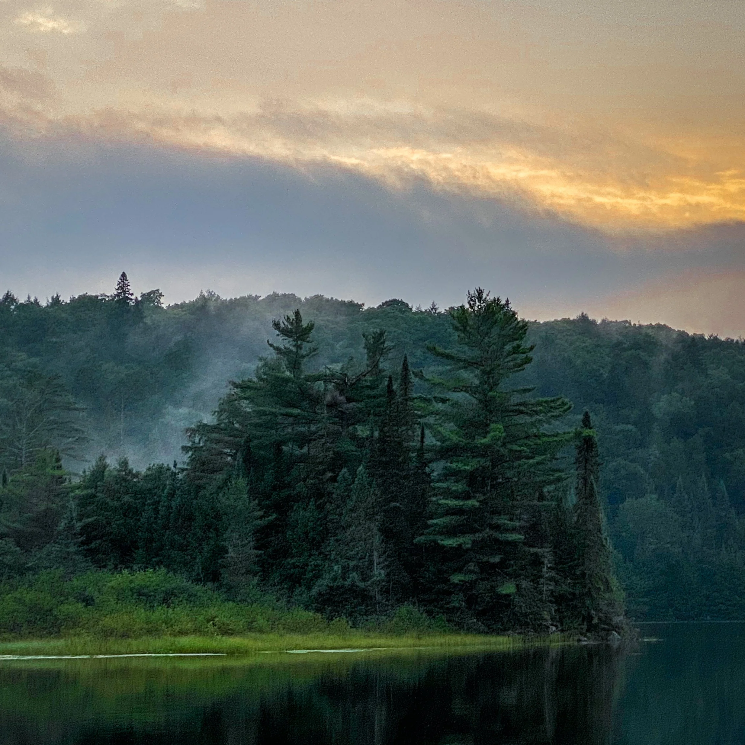 Sunset over a forested lake with trees reflected in the water and a sky with orange and gray clouds.