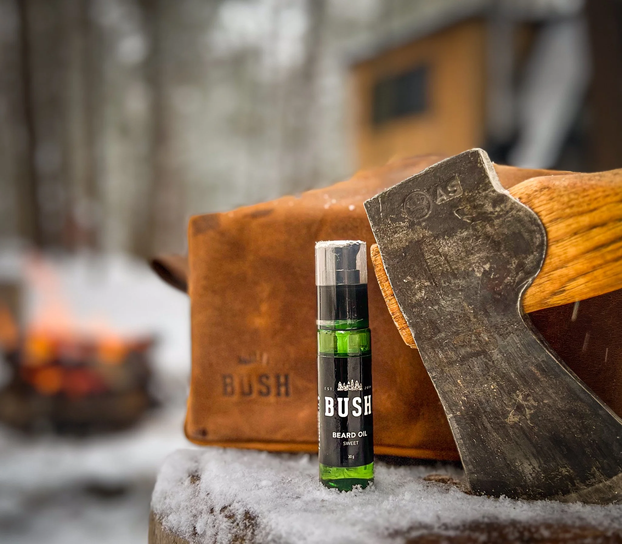 Small jar of BUSH beard oil placed on a wooden surface outdoors, with an axe and a leather carrying case in the background.