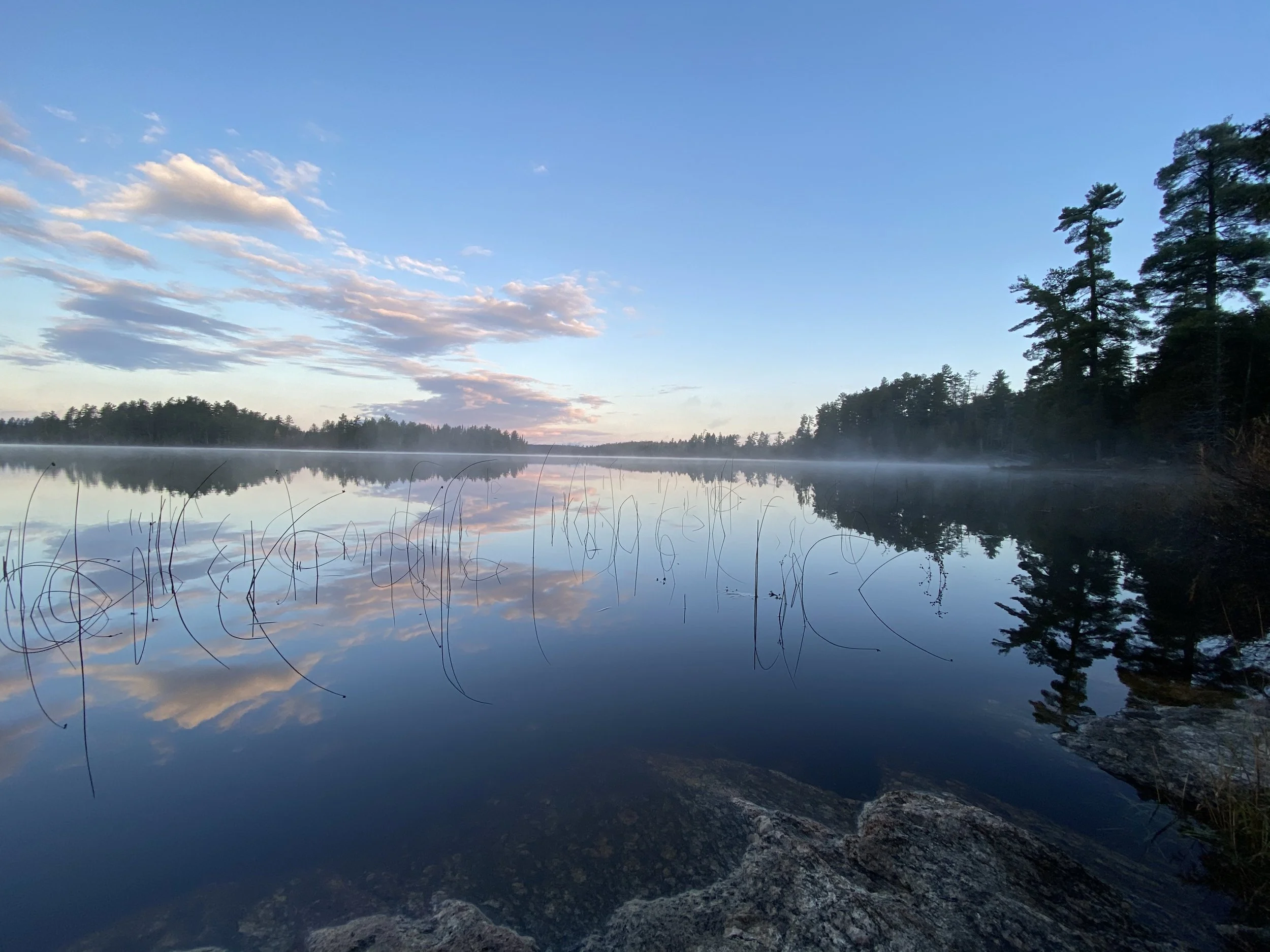 A calm lake reflecting a partly cloudy sky during sunrise or sunset, with trees on the far shoreline and rocks in the foreground.
