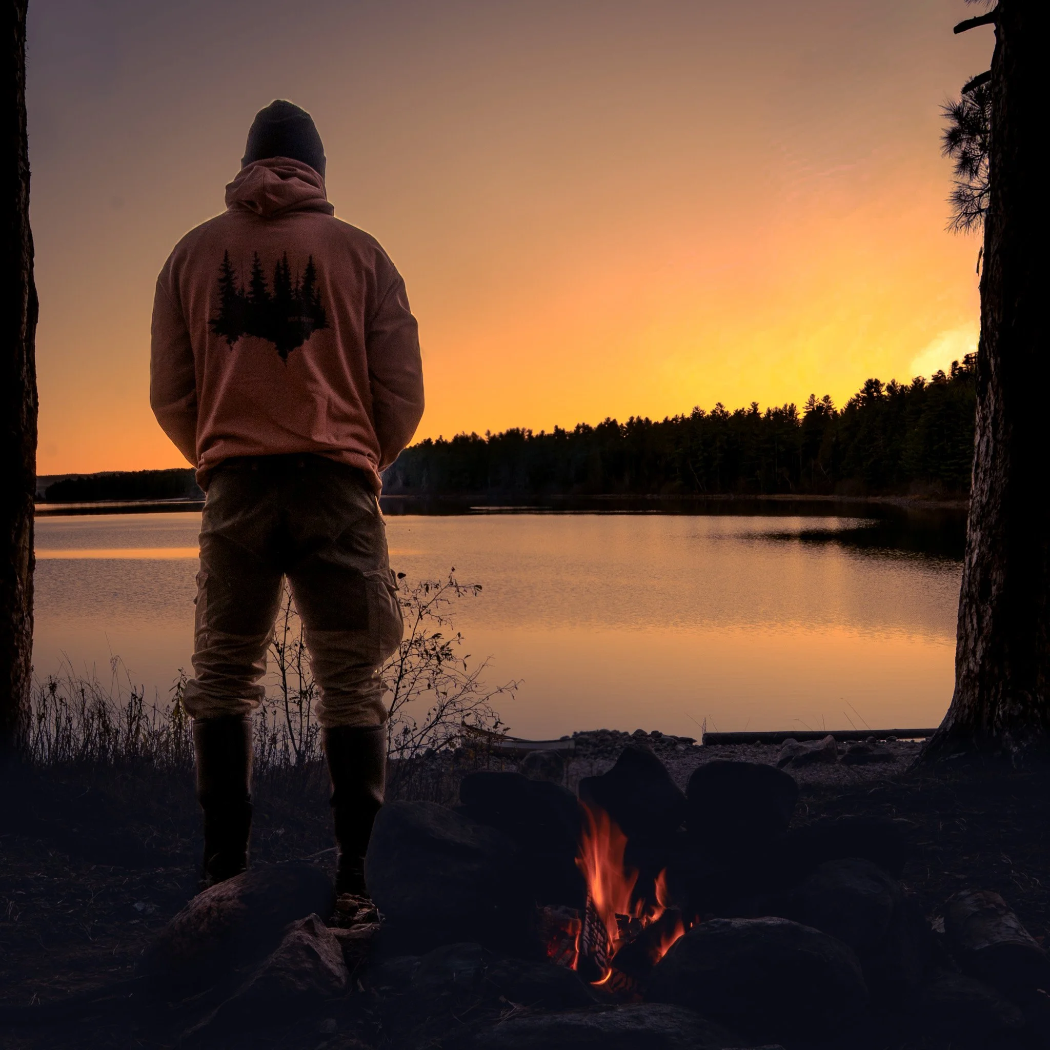Person standing by a campfire looking at a sunset over a lake, surrounded by trees.