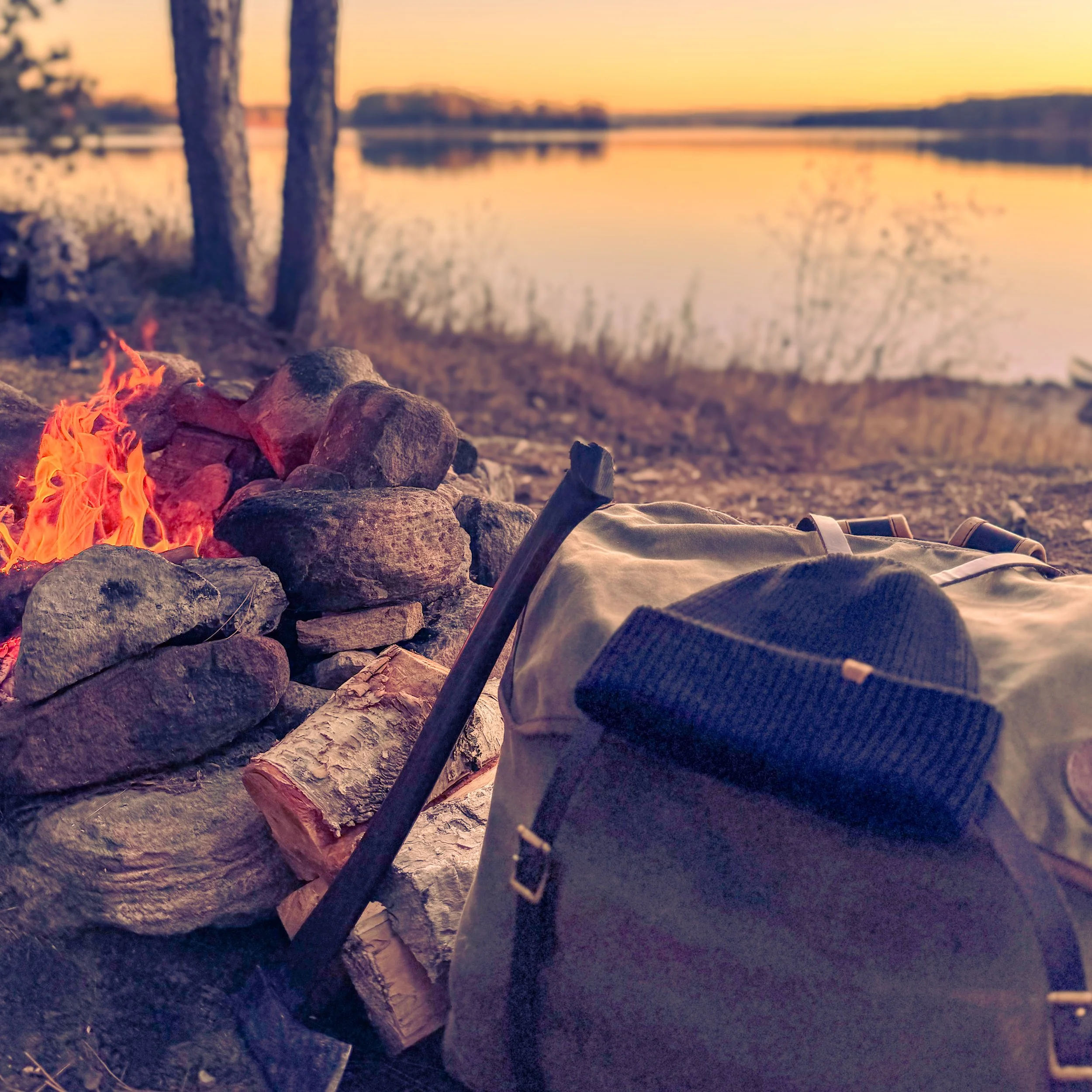 Campfire by a lake at sunset with a backpack and walking stick in the foreground.