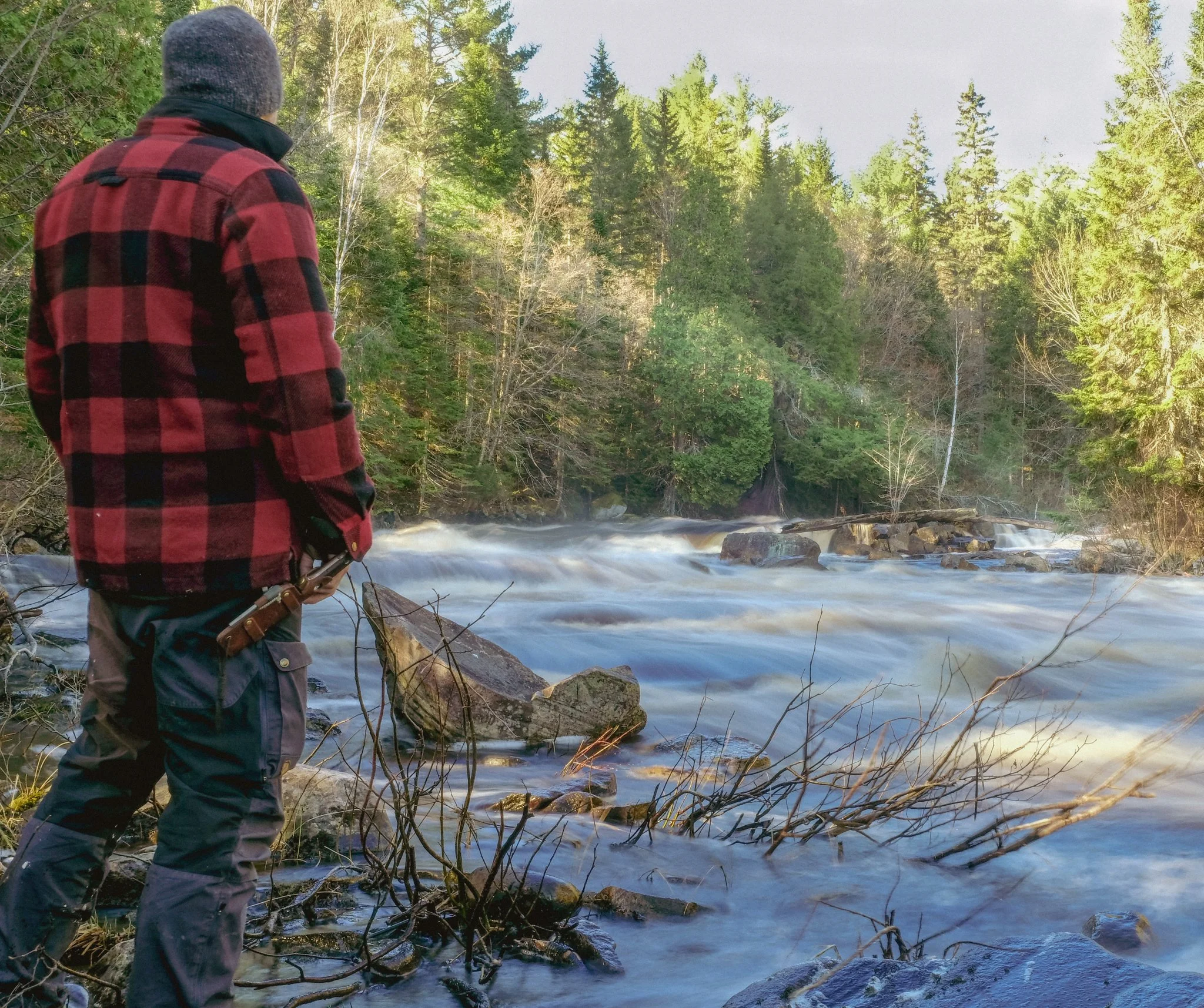 A person wearing a red and black checkered jacket, gray beanie, and outdoor gear standing by a flowing river in a forest with green trees.