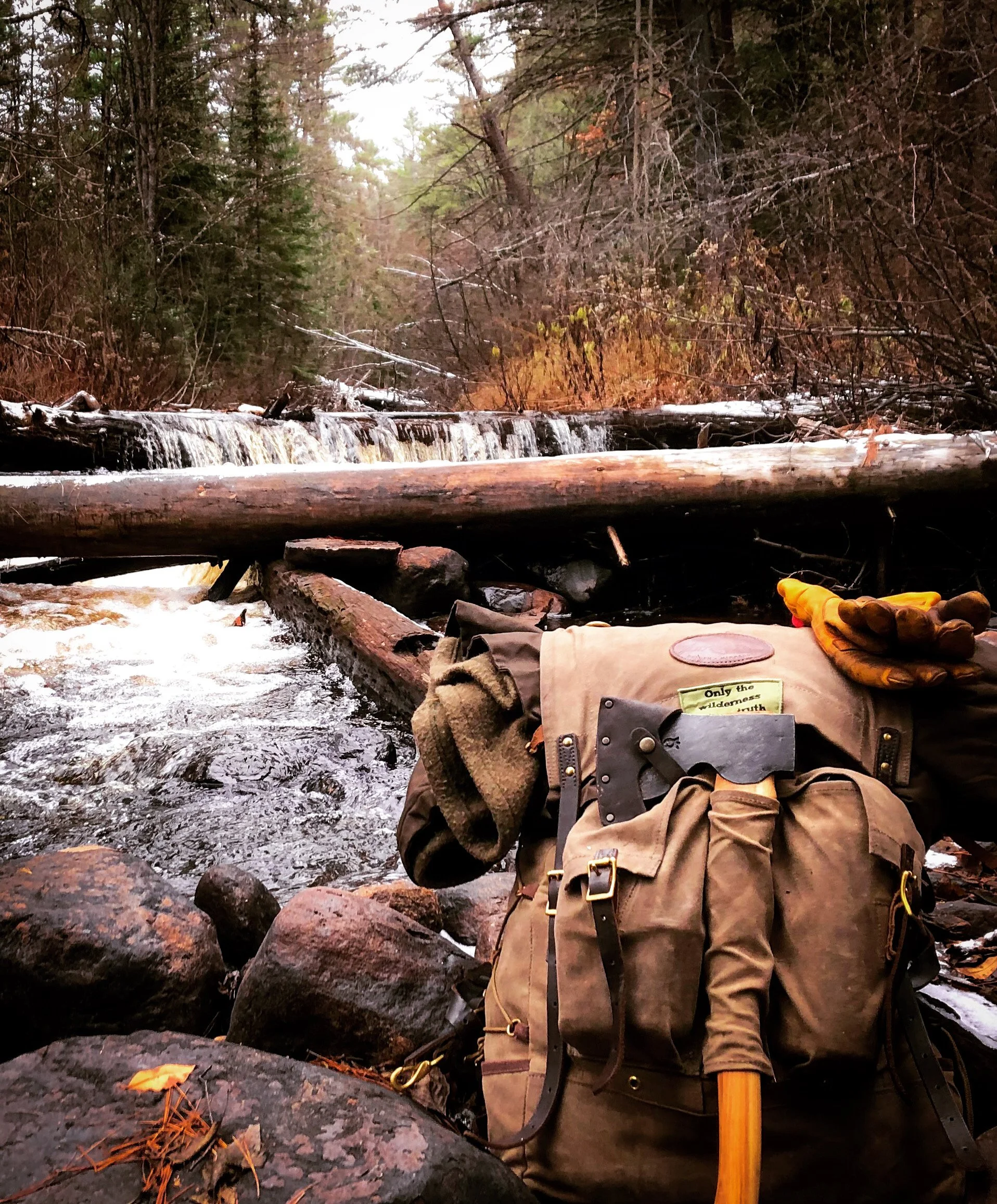 A brown hiking backpack with gear on rocks beside a small stream and a fallen tree, in a wooded forest area.