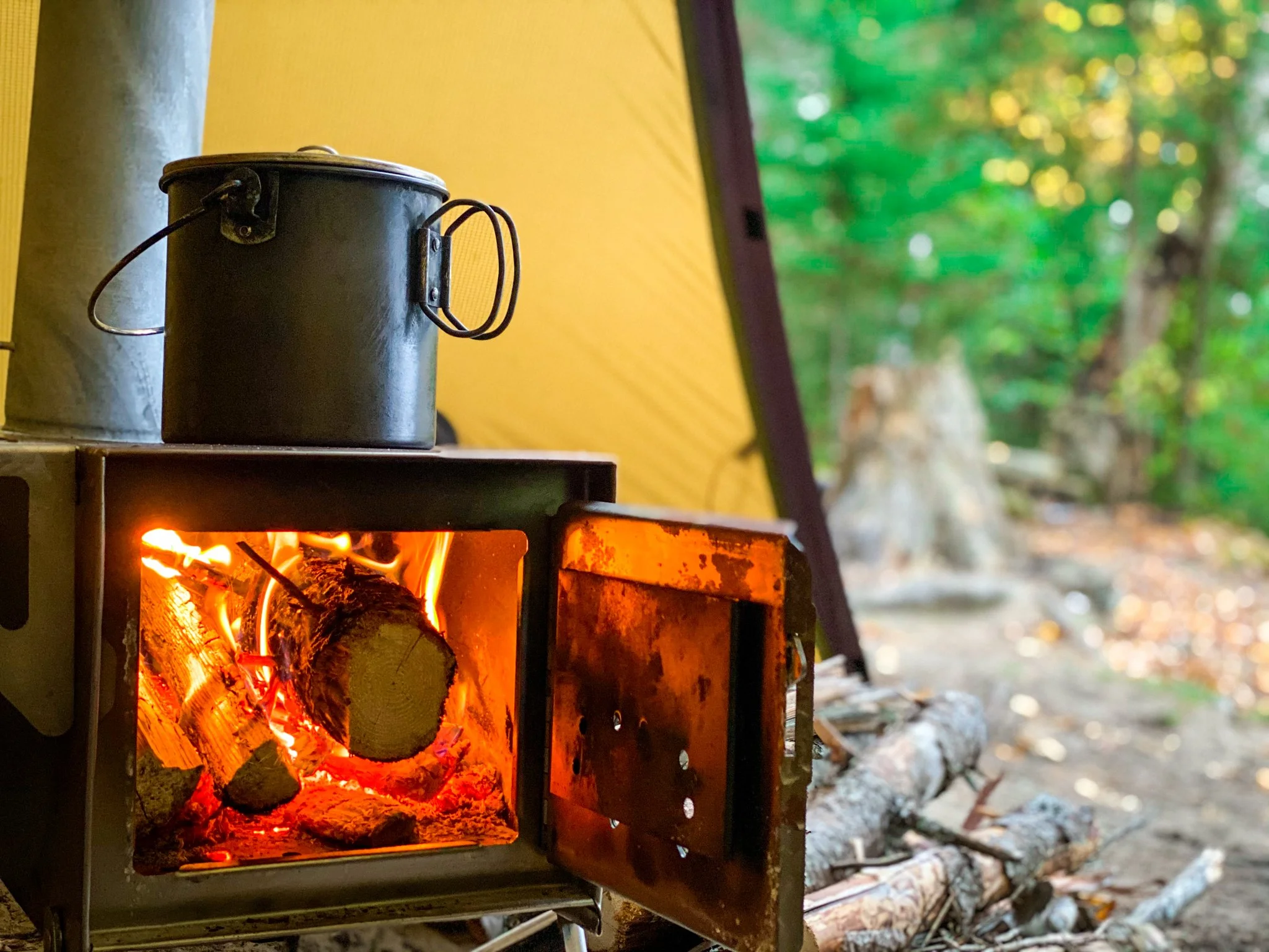 A small metal camping stove with a lit fire inside, with logs and flames visible, placed outside near a yellow tent and surrounded by trees.