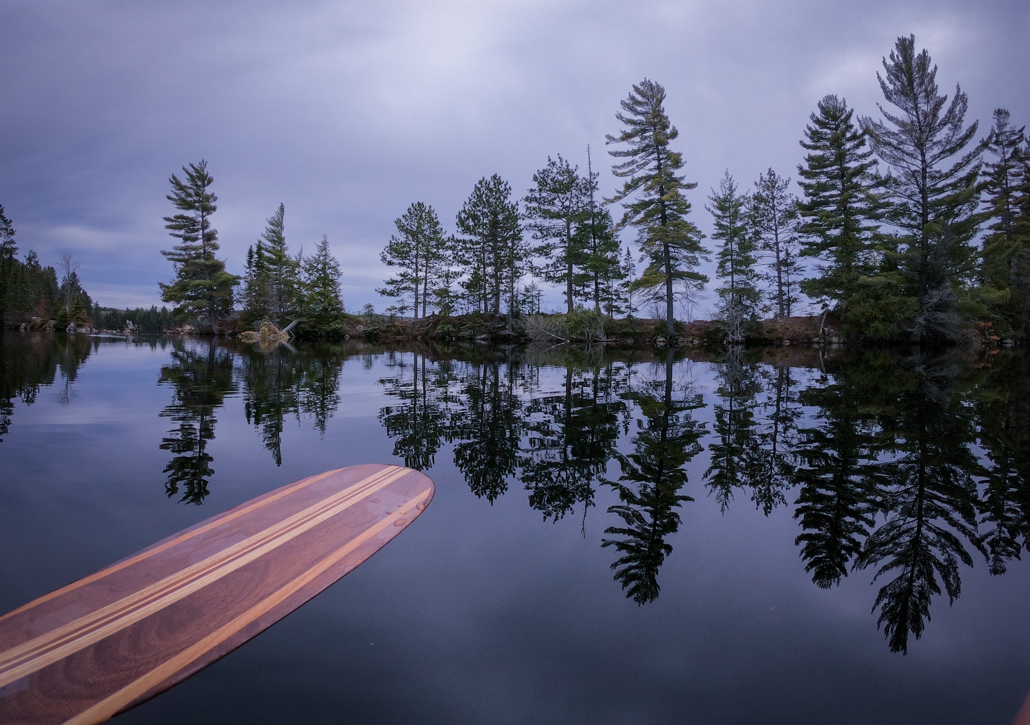 View of a lake with calm water reflecting surrounding pine trees and cloudy sky, with a wooden paddle partially visible in the foreground.