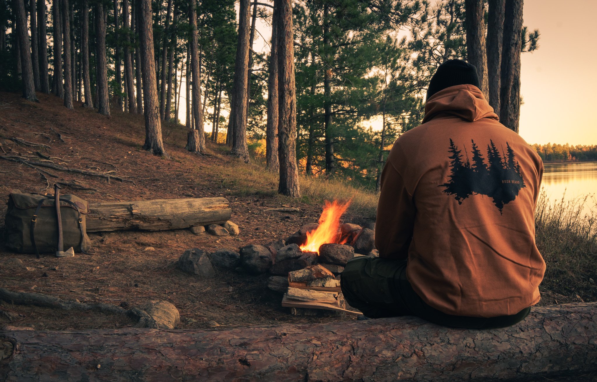 Person sitting by a campfire in a forest near a lake at sunset, wearing a brown jacket with a tree graphic on the back