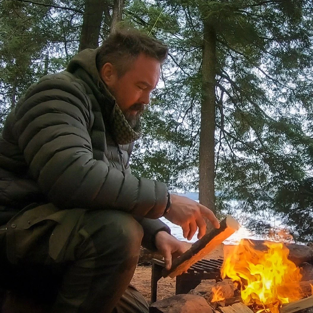 Man wearing a dark puffer jacket cooking over an outdoor fire in a wooded area.