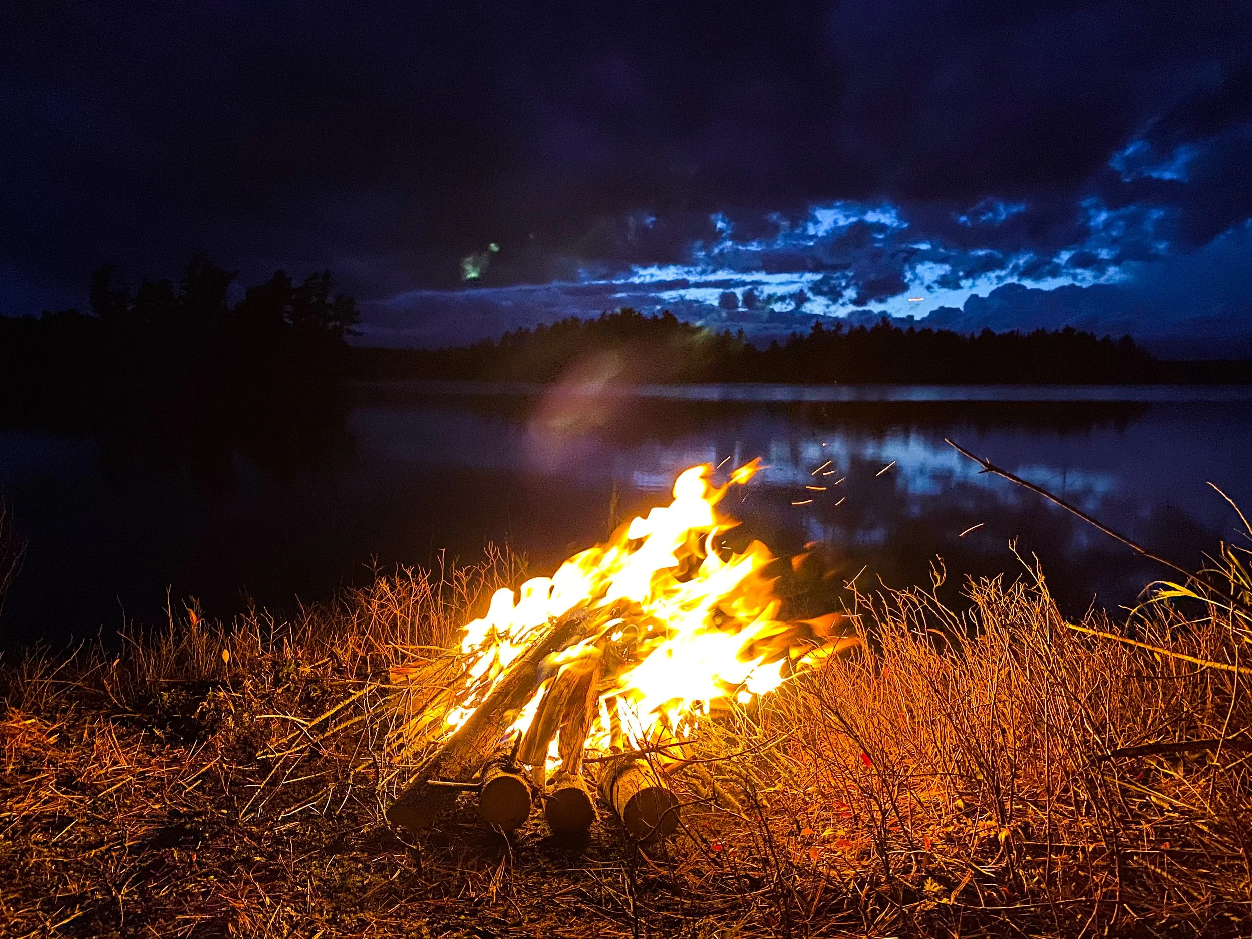 Campfire burning near a body of water under a dark, cloudy night sky.