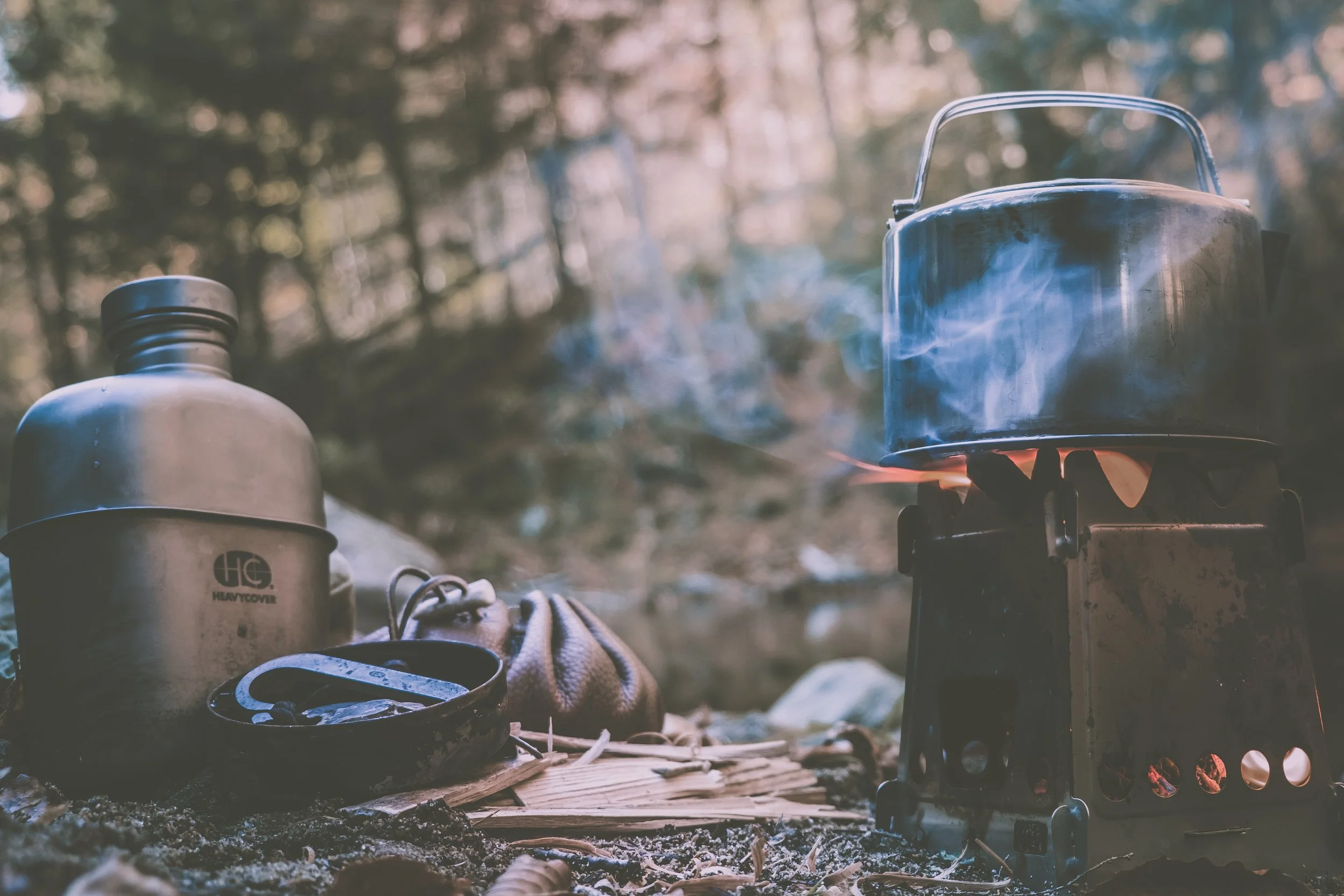 Camping stove with something cooking, an outdoor canister, and a cup placed on a wooden surface in a forested outdoor setting.
