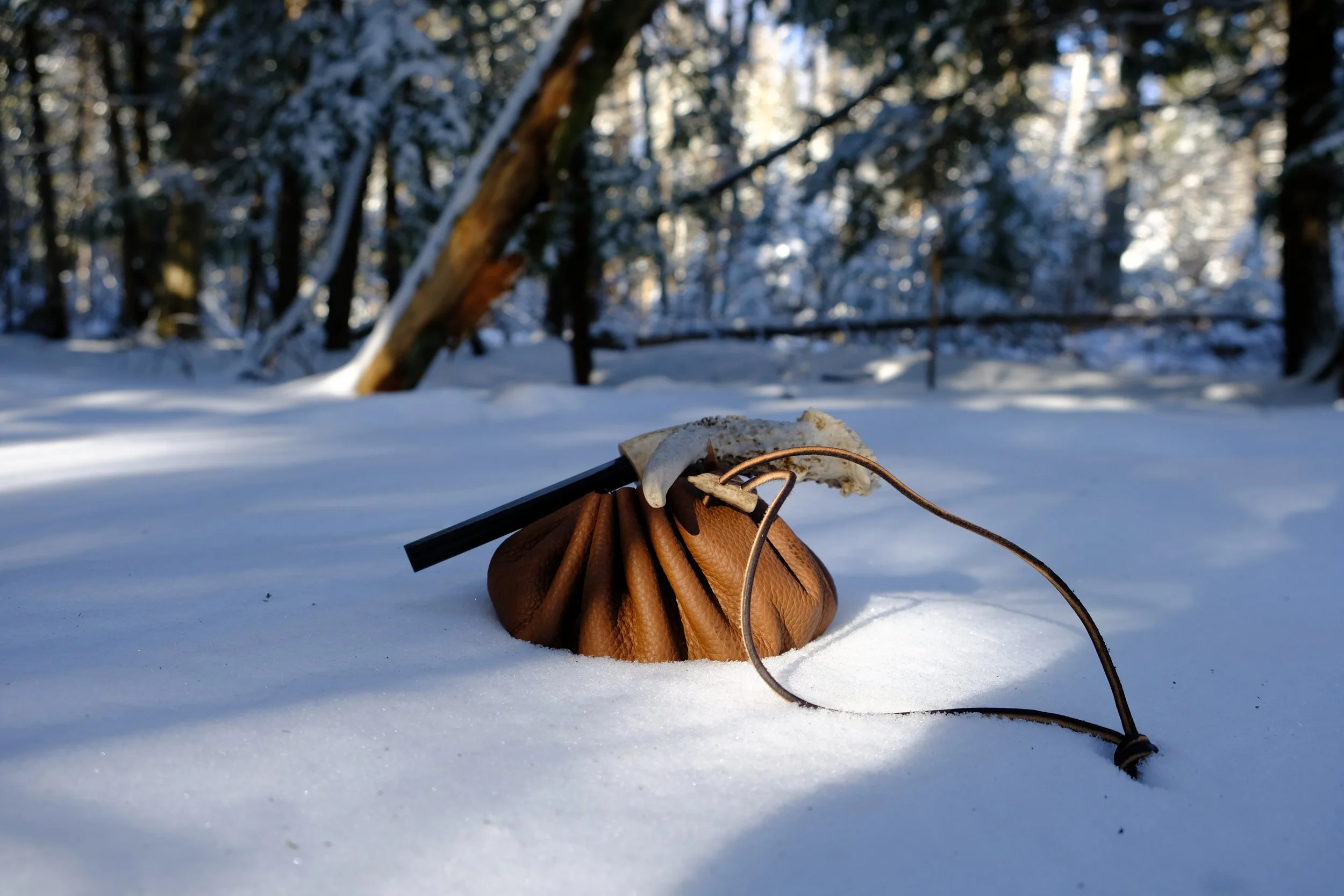 A brown leather drawstring bag on snow in a forest with trees and sunlight in the background.