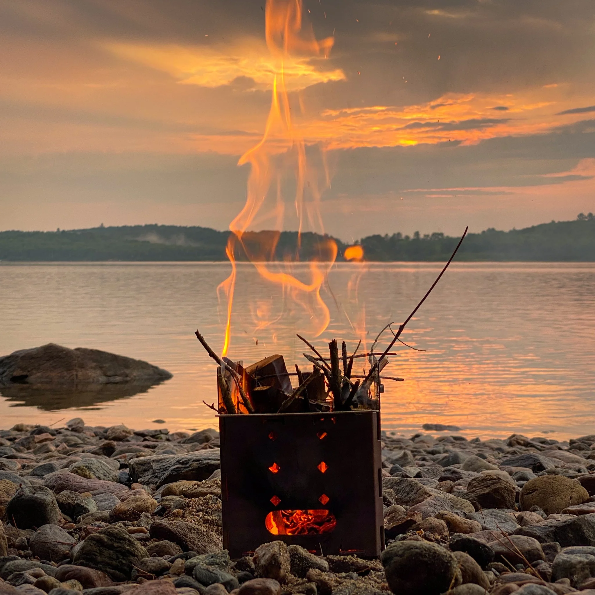 A small portable fire pit with sticks burning inside, set on a rocky lakeshore during sunset, with water, a rock, and distant trees in the background.