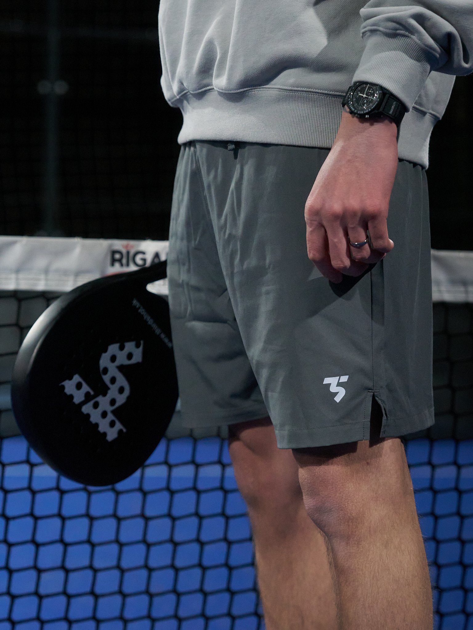 A person wearing grey athletic shorts and a grey sweatshirt, holding a black pickleball paddle with a white logo, standing near a blue pickleball court net.