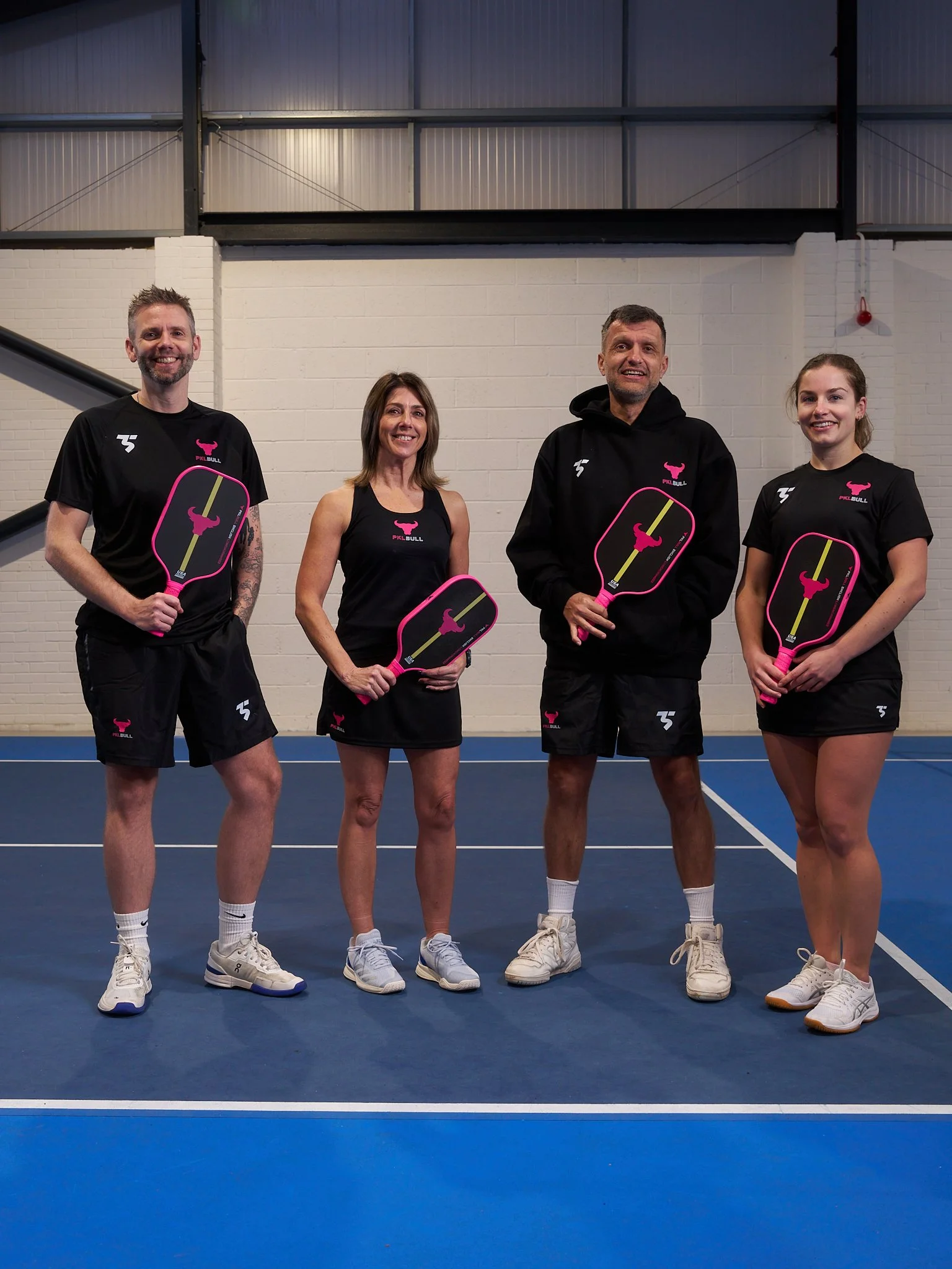 Four people standing on a blue basketball court, holding pink paddles with a pink bull logo, wearing black sports apparel with pink logos, smiling at the camera.