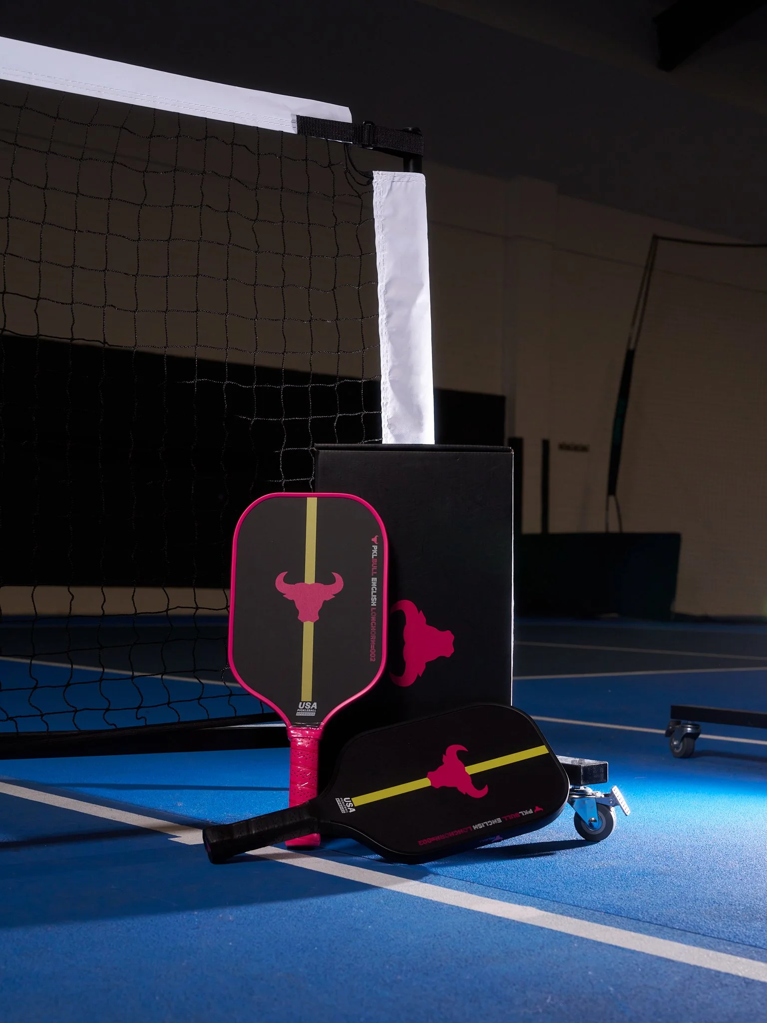 Two pickleball paddles resting on a blue court next to a black box, with a pickleball net in the background.