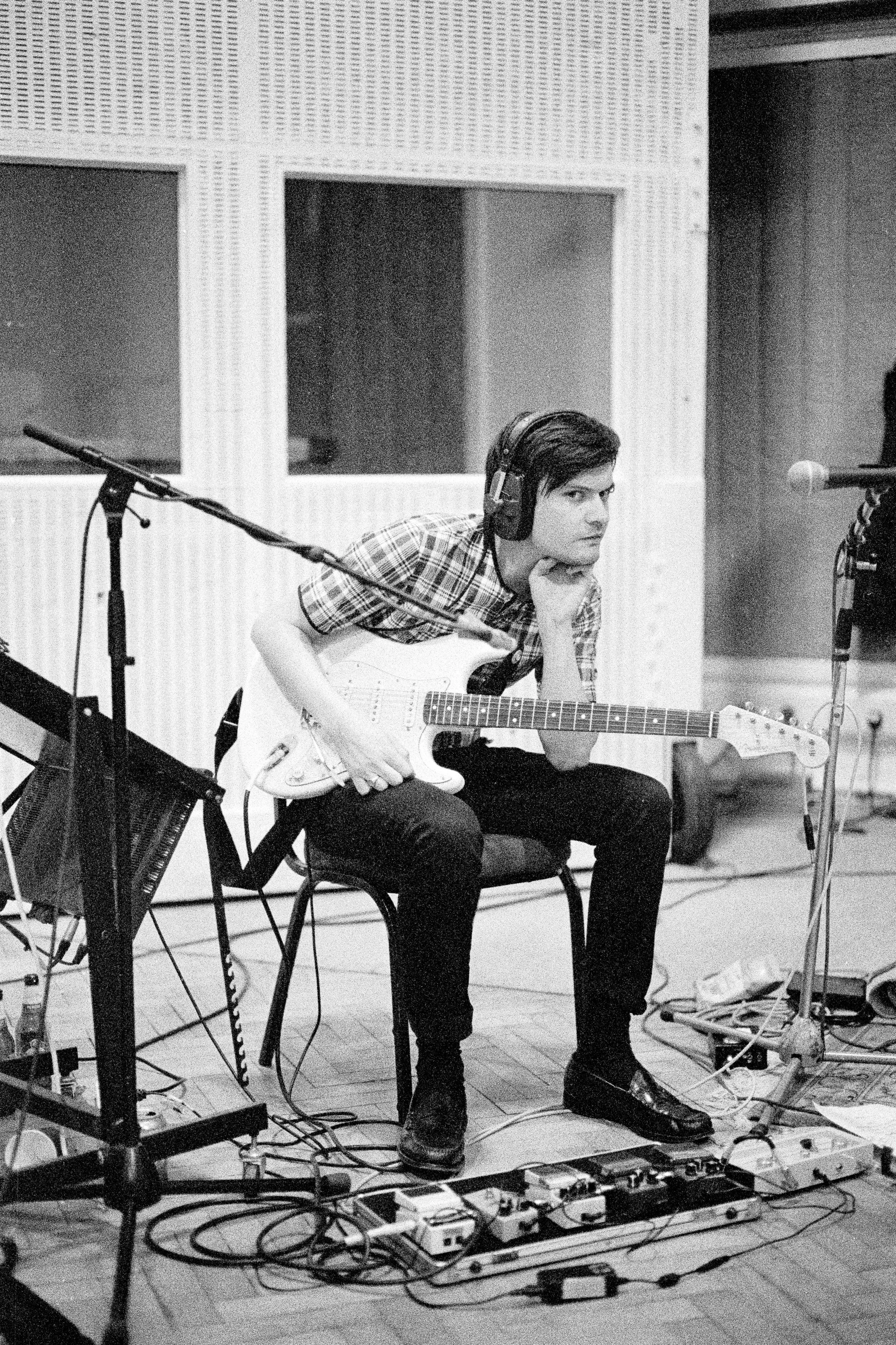 A young man with dark hair wearing a checkered shirt, sitting in a recording studio with an electric guitar, headphones, and various audio equipment.
