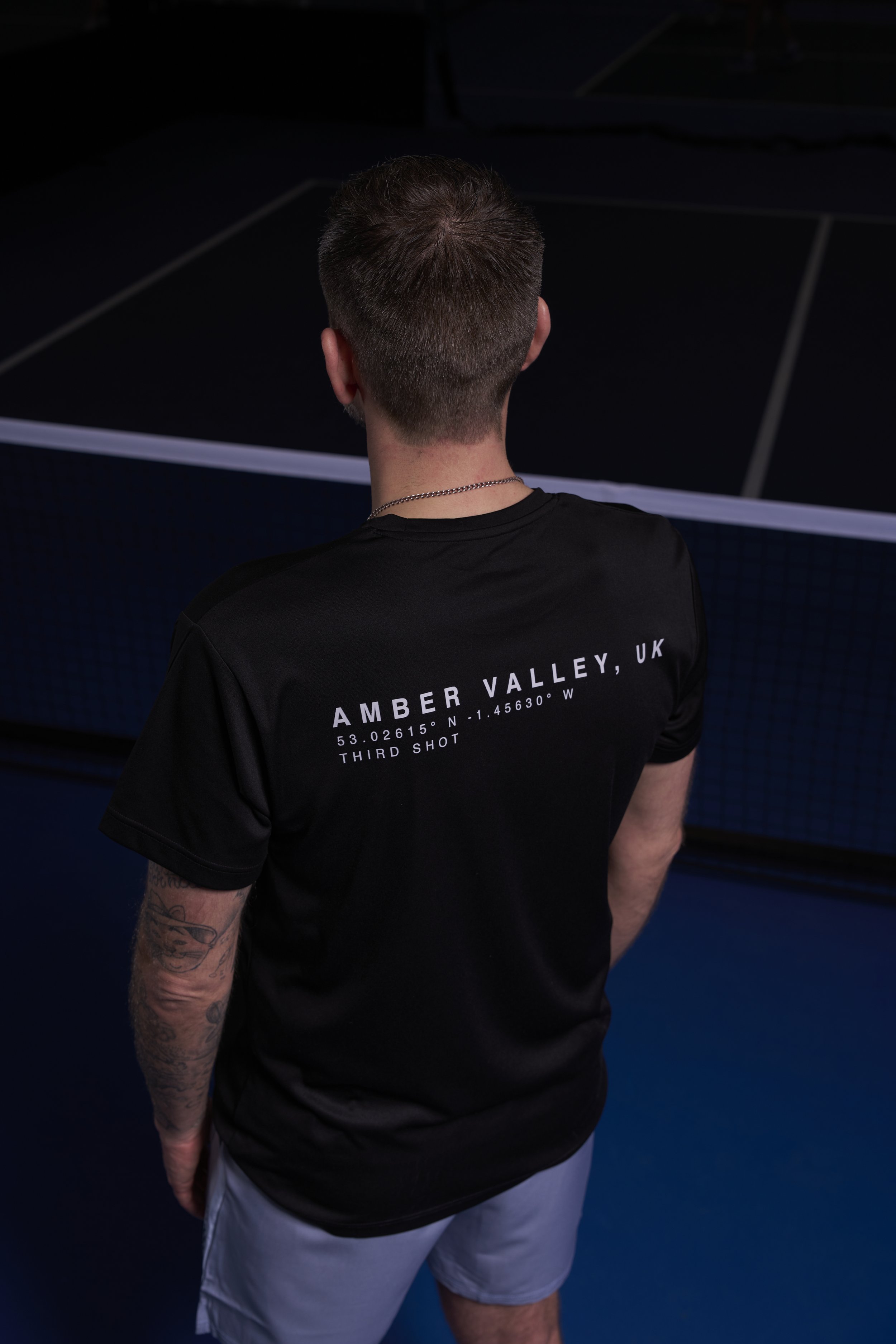 Back of a young man standing in front of a ping pong table, wearing a black T-shirt with white text about Amber Valley, UK, and the phrase 'Third Shot,' with tattoos on his left arm and short, dark hair.