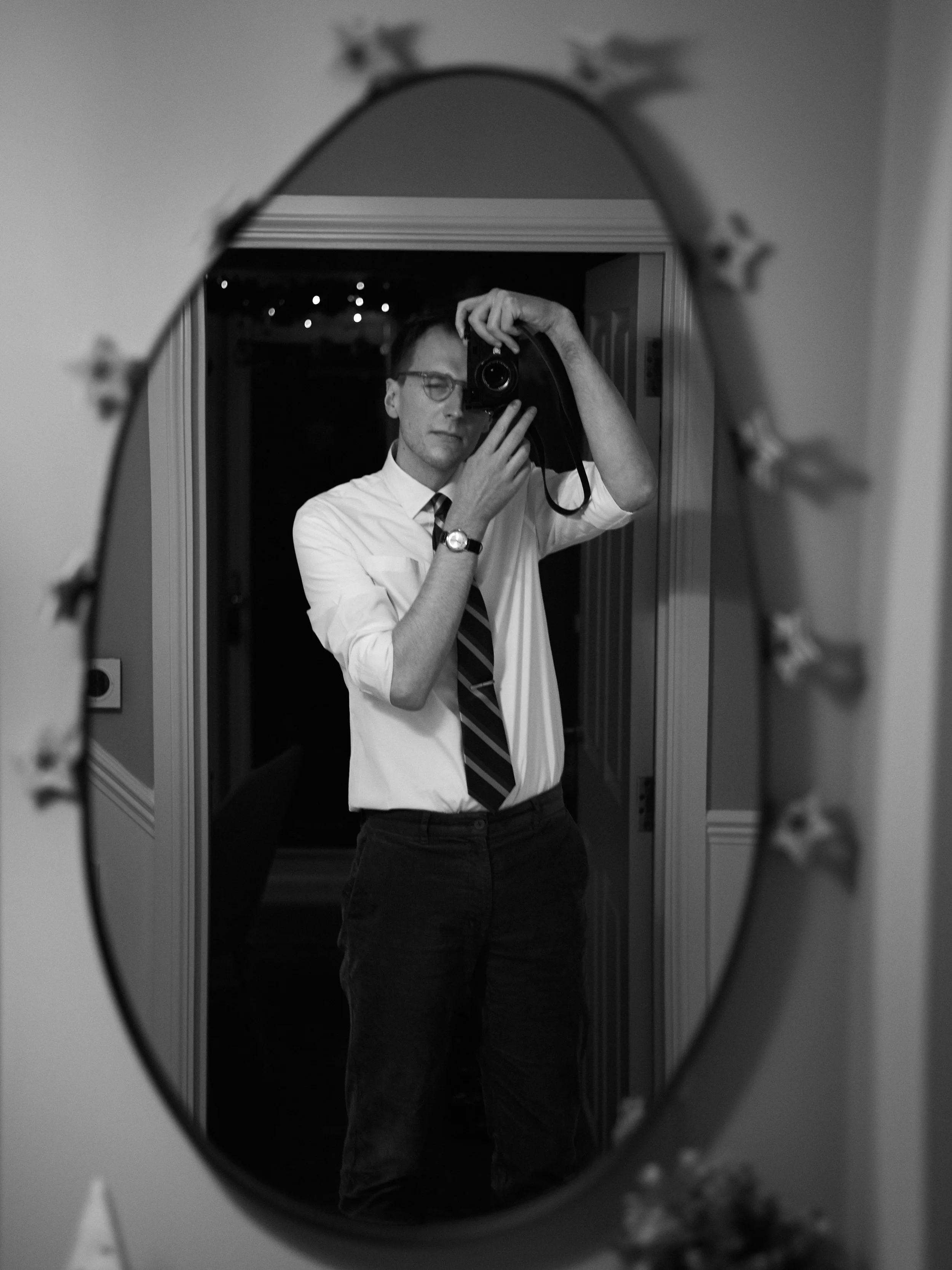 A man is taking a mirror selfie in a hallway, wearing glasses, a white shirt, a striped tie, and dark pants, holding a camera up to his face.