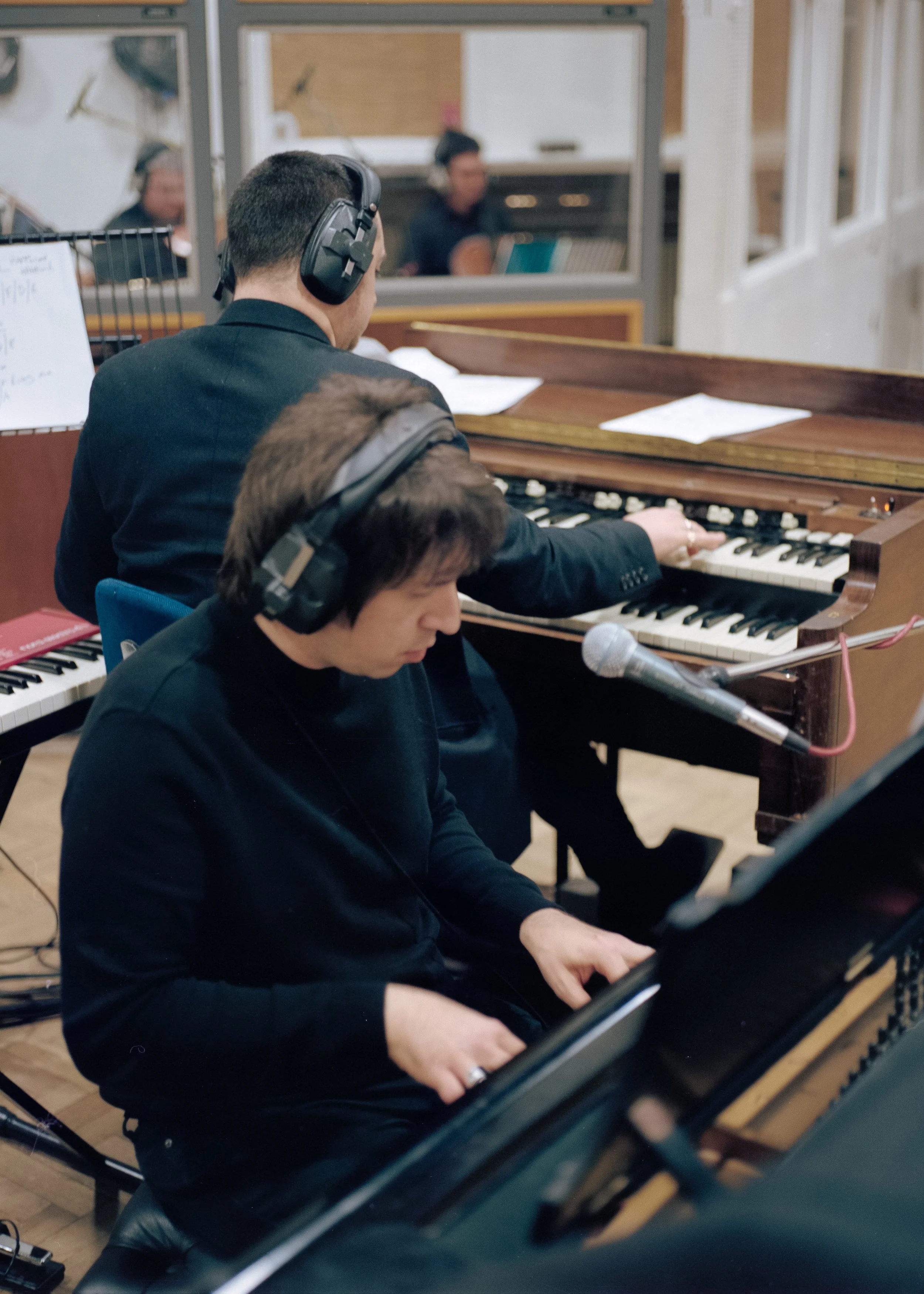 Two musicians with headsets playing pianos in a studio, with a woman visible in the mirror behind them.