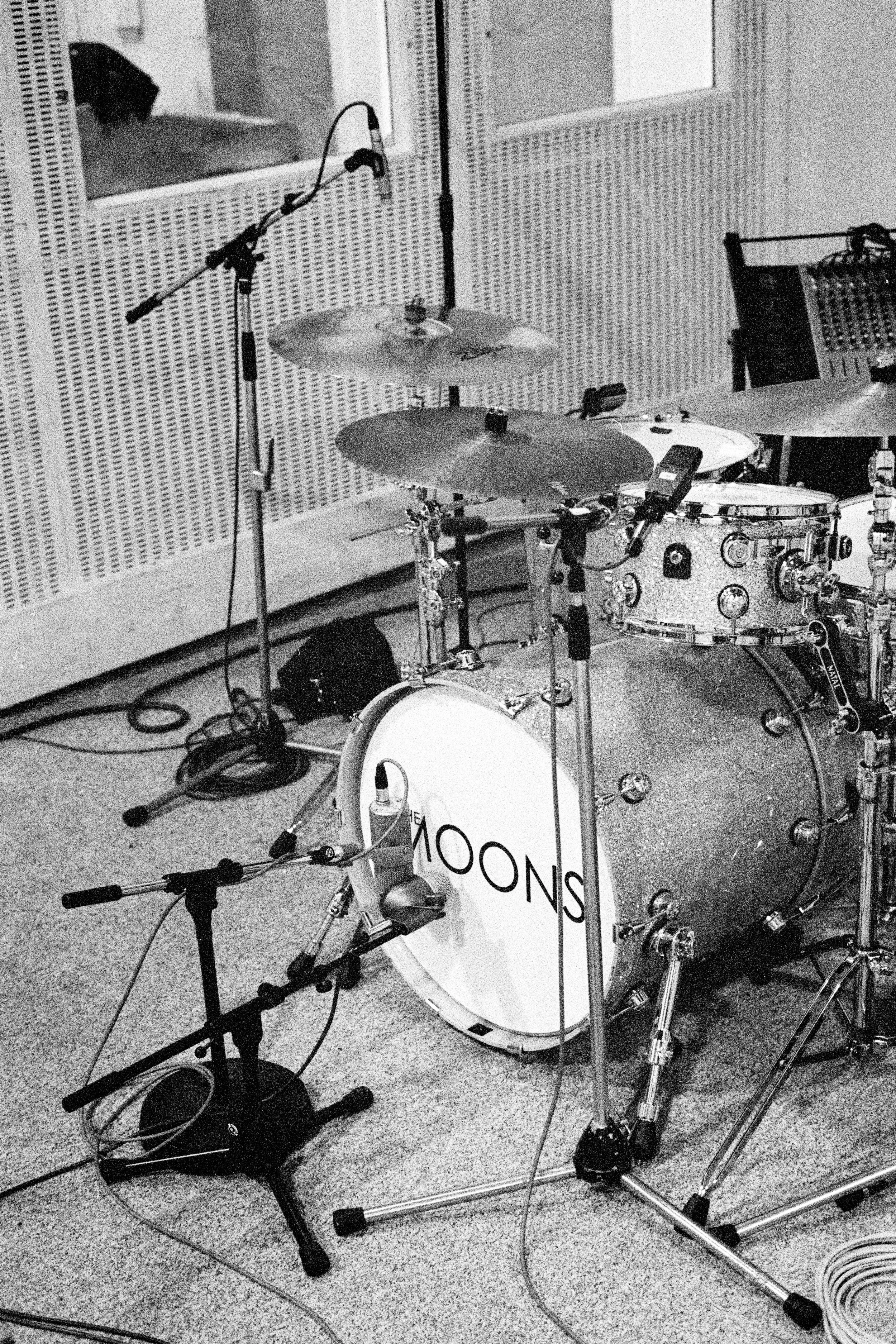 A drum set with a bass drum labeled 'MOONS,' cymbals, and drums in a music recording studio, with microphones positioned around and electrical cables on the floor.